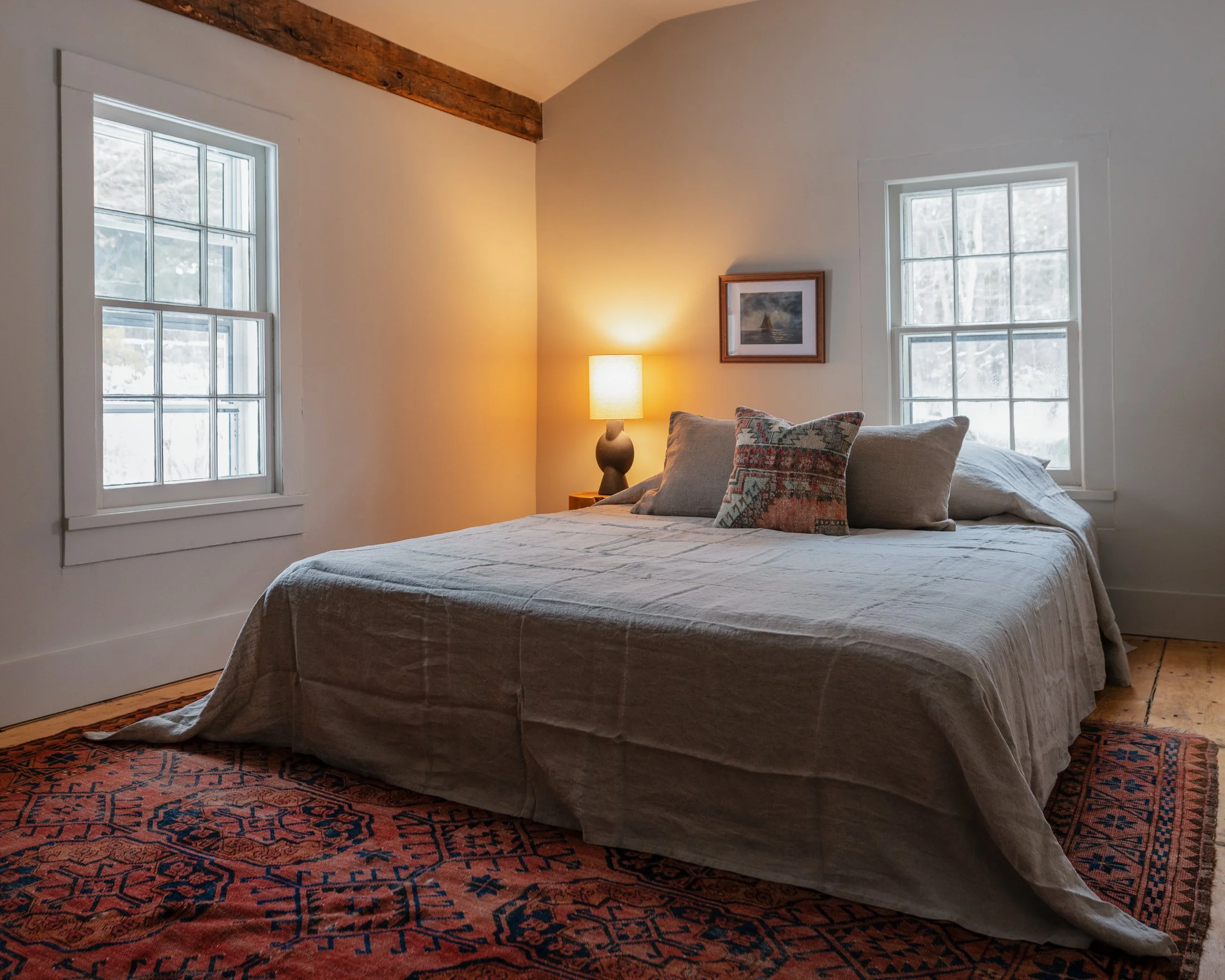 A cozy bedroom with a large bed draped in beige linen, accented by decorative pillows. There is a warm table lamp on a nightstand and two windows with white trim letting in natural light. The room has a wooden floor and an orange patterned rug, and a