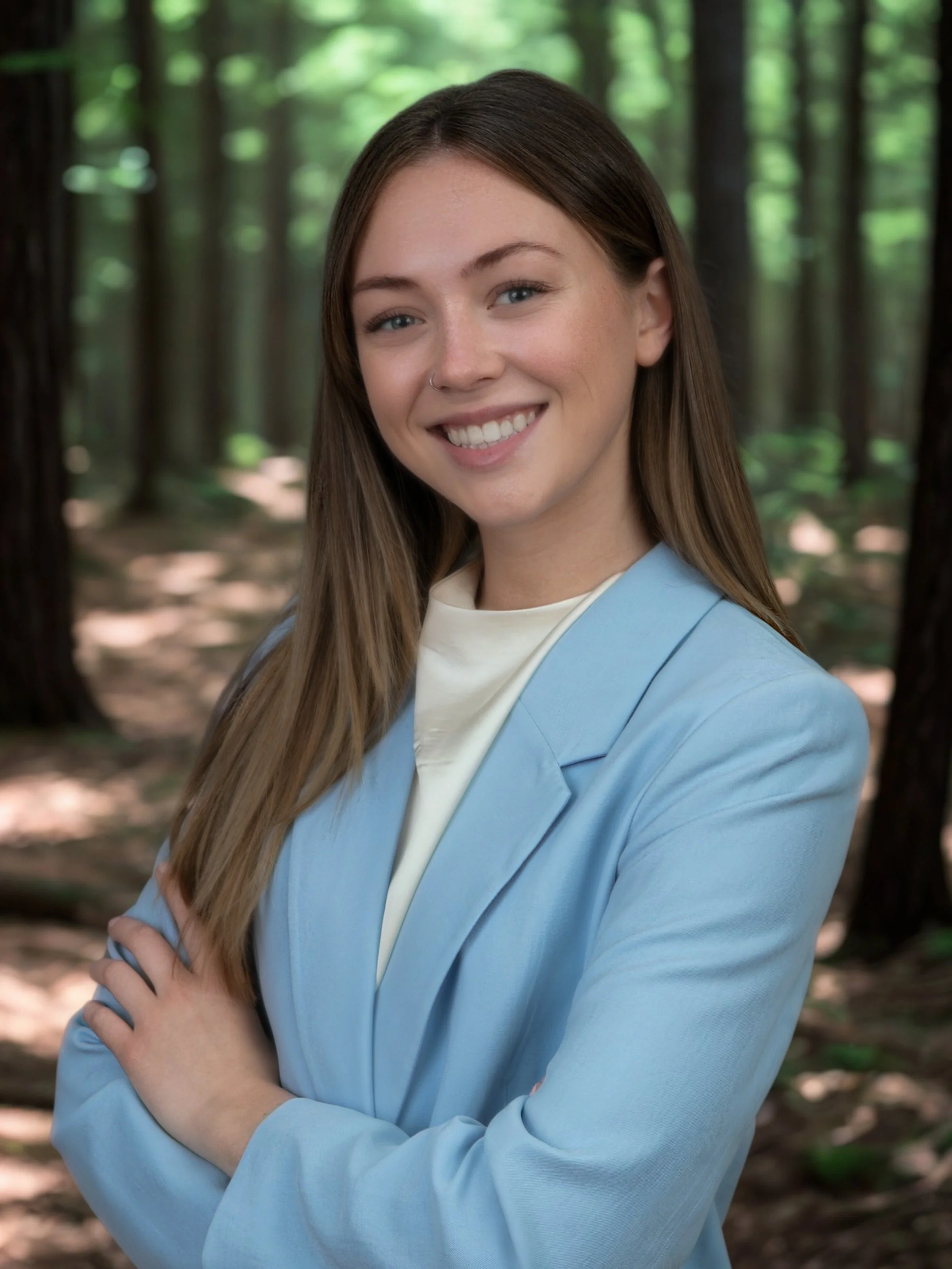 A woman with long brown hair, blue eyes, and a nose piercing, smiling and standing in a forest, wearing a light blue blazer over a white shirt.
