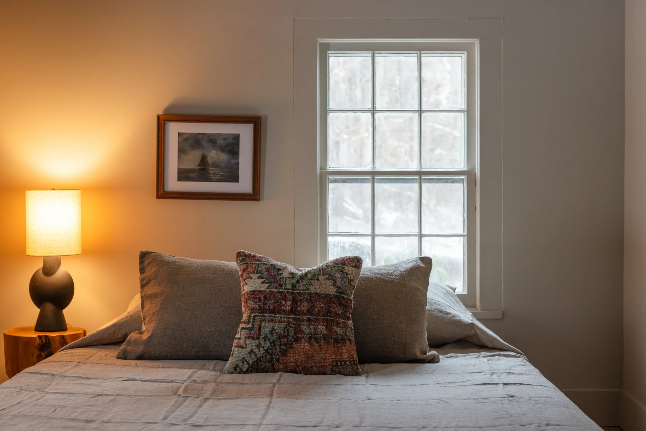 A cozy bedroom with a bed featuring beige bedding, three pillows including a patterned decorative pillow, a wooden nightstand with a lamp, a framed picture on the wall, and a window showing a snowy outdoor scene.