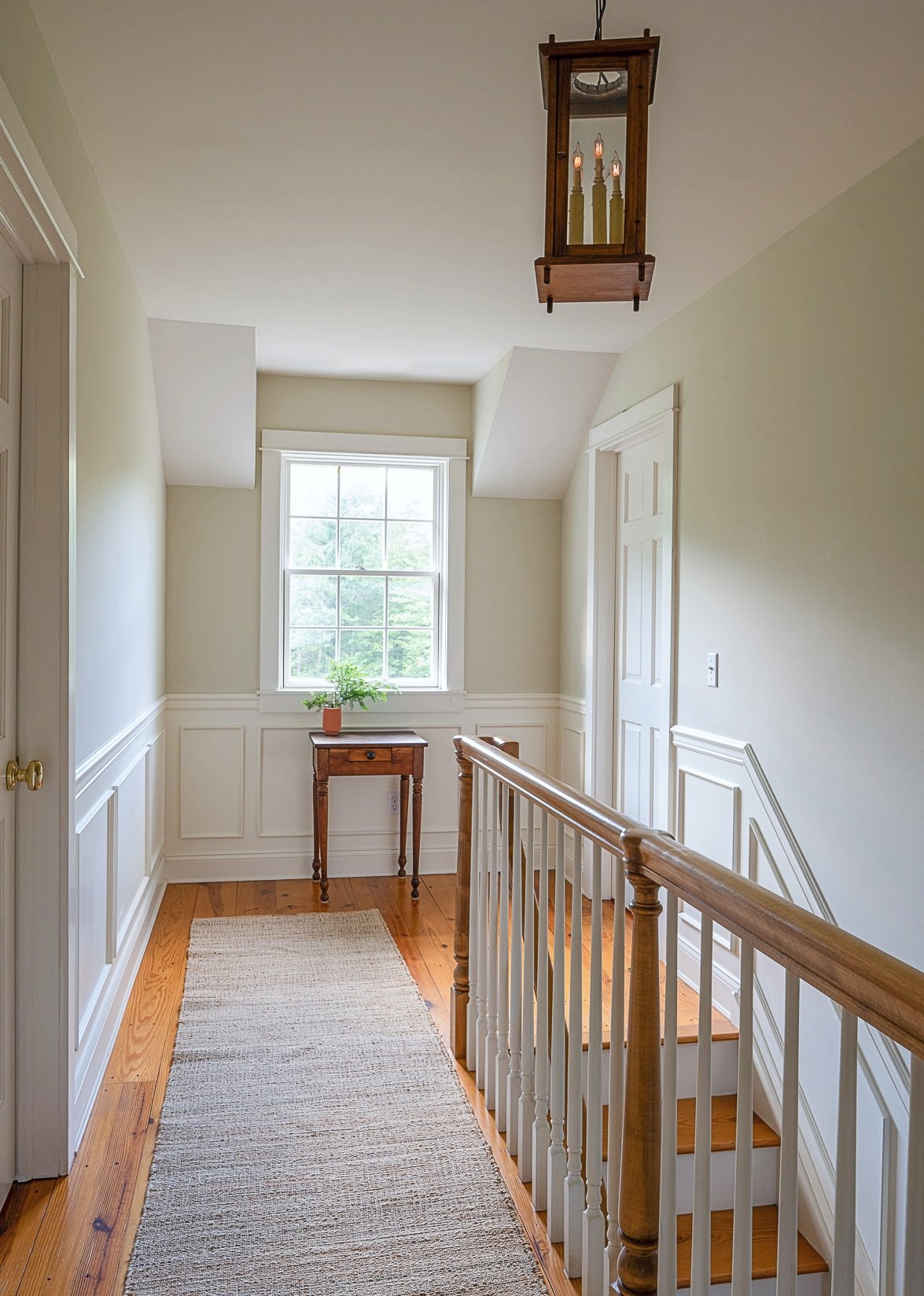 A hallway landing with a window, a small wooden table with a potted plant, a hanging wooden lantern-style light fixture, a staircase with wooden handrails and white spindles, oak flooring, and wainscoting on the walls.