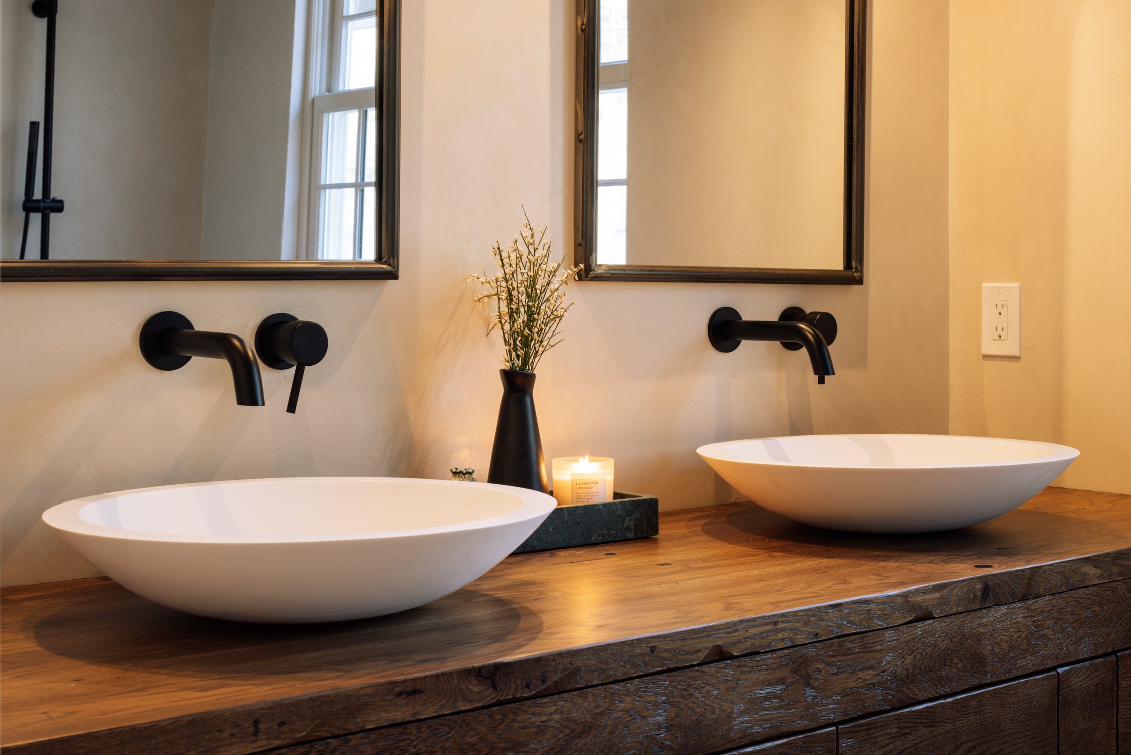 A bathroom with two white vessel sinks on a rustic wooden countertop, black wall-mounted faucets, two mirrors, a black vase with greenery, a lit candle, and a double electrical outlet.