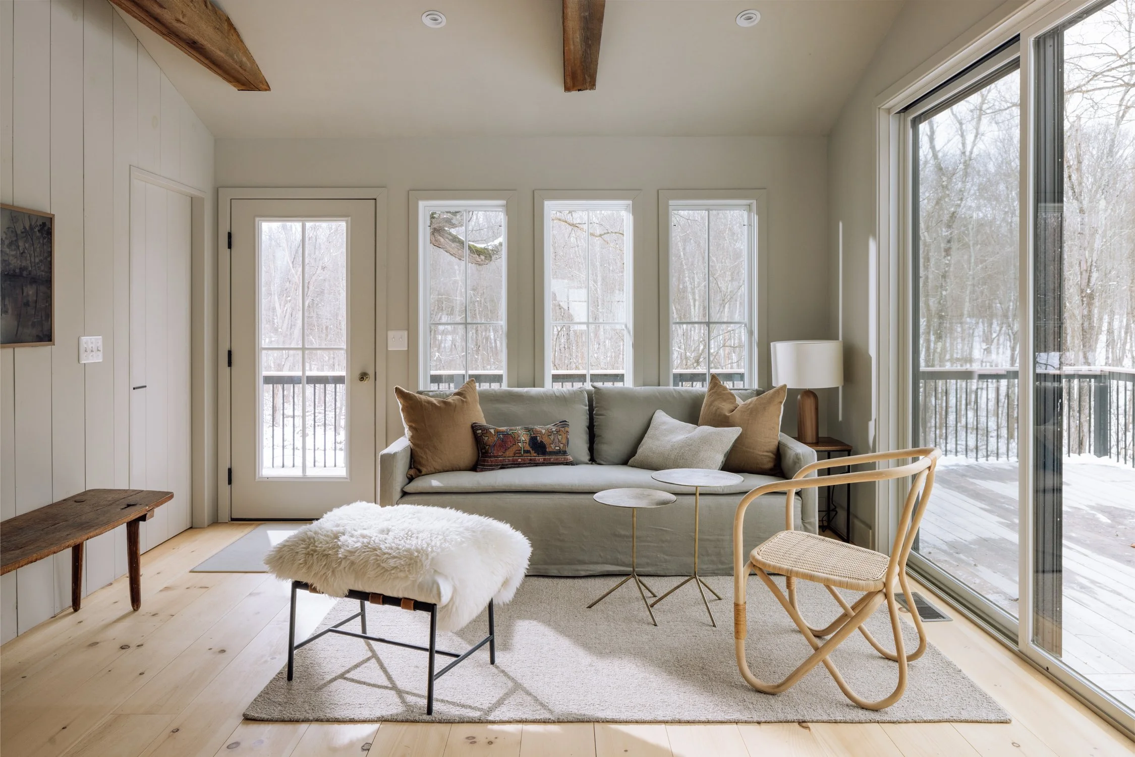 Living room with light-colored walls, a sofa with multiple pillows, a side table with a lamp, a rattan chair, a fluffy stool, and large windows showing a snowy outdoor scene.