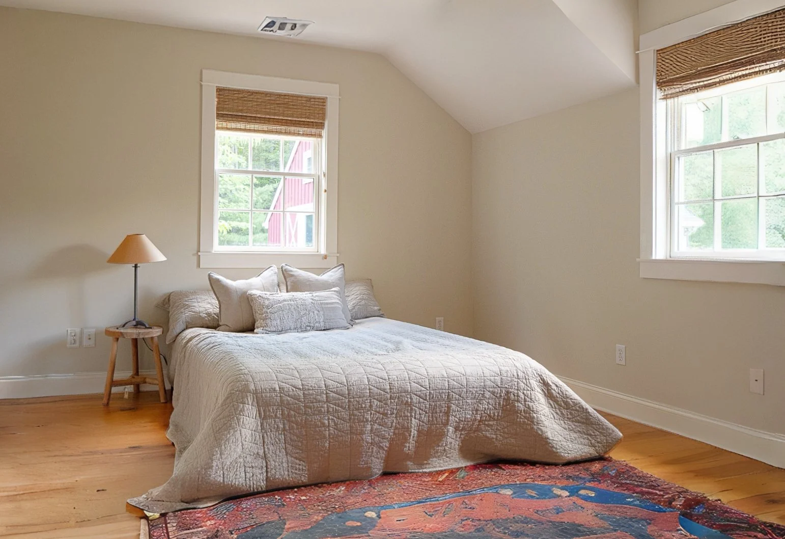 A bedroom with a bed covered in a light-colored quilt, several pillows, a wooden nightstand with a lamp, two windows with blinds, hardwood flooring, and a colorful area rug.