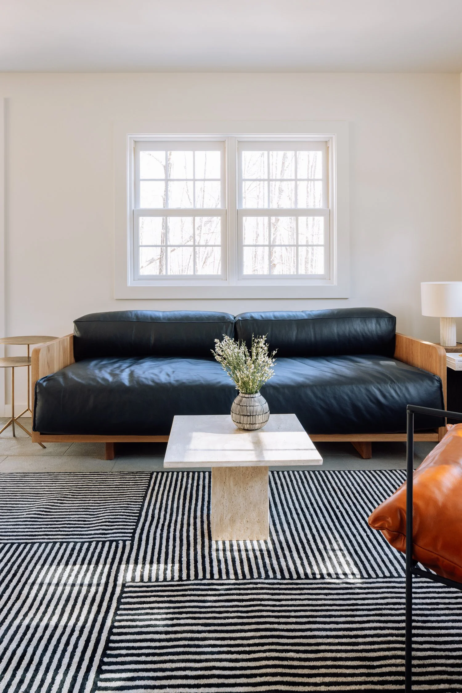 Living room with a black leather sofa, a small marble coffee table with a vase of white flowers, a black and white rug, a side table, and a window with frosted glass letting in natural light.
