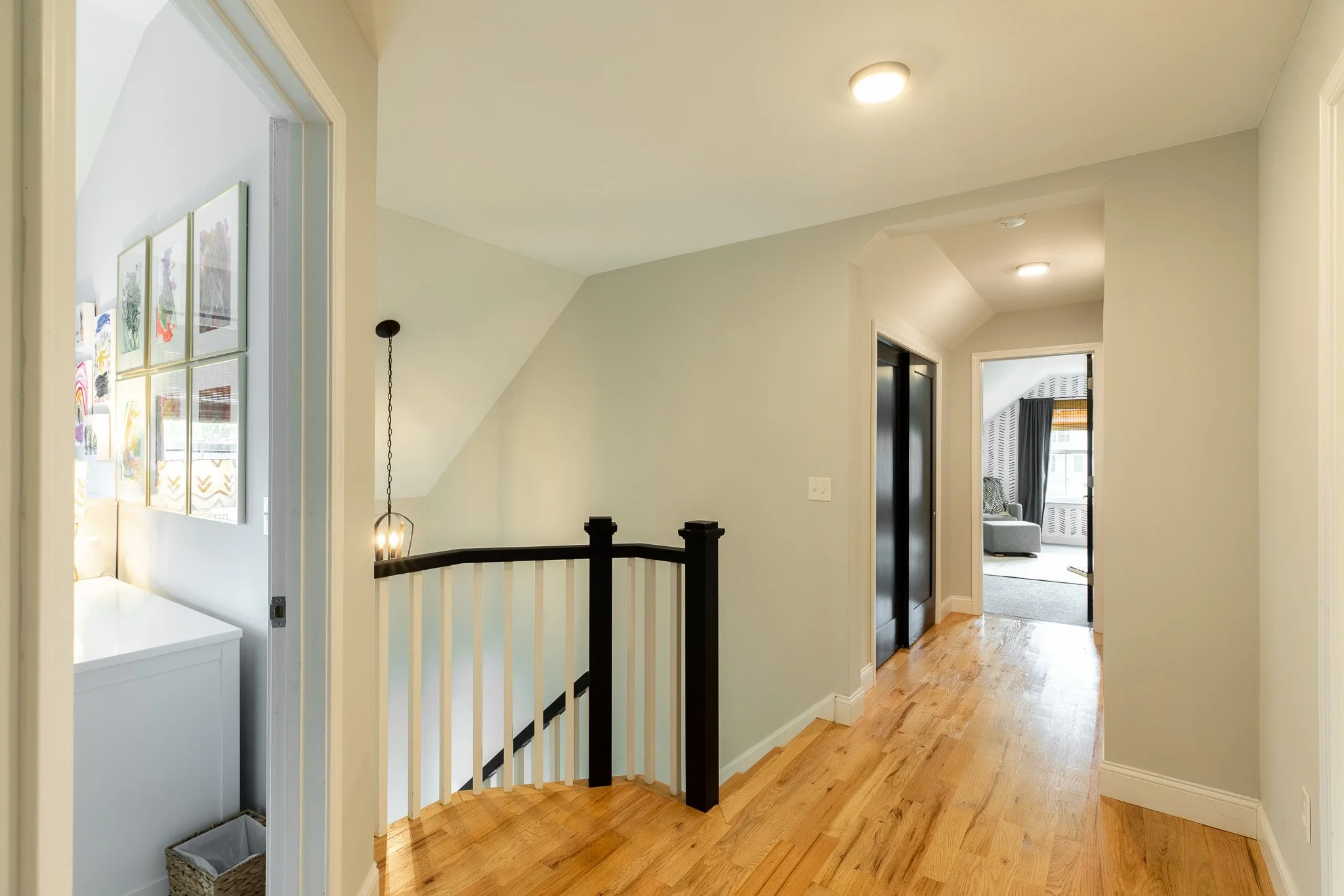 Hallway with light-colored walls, wooden flooring, an elevator with black doors, and visible rooms with artwork and furniture.