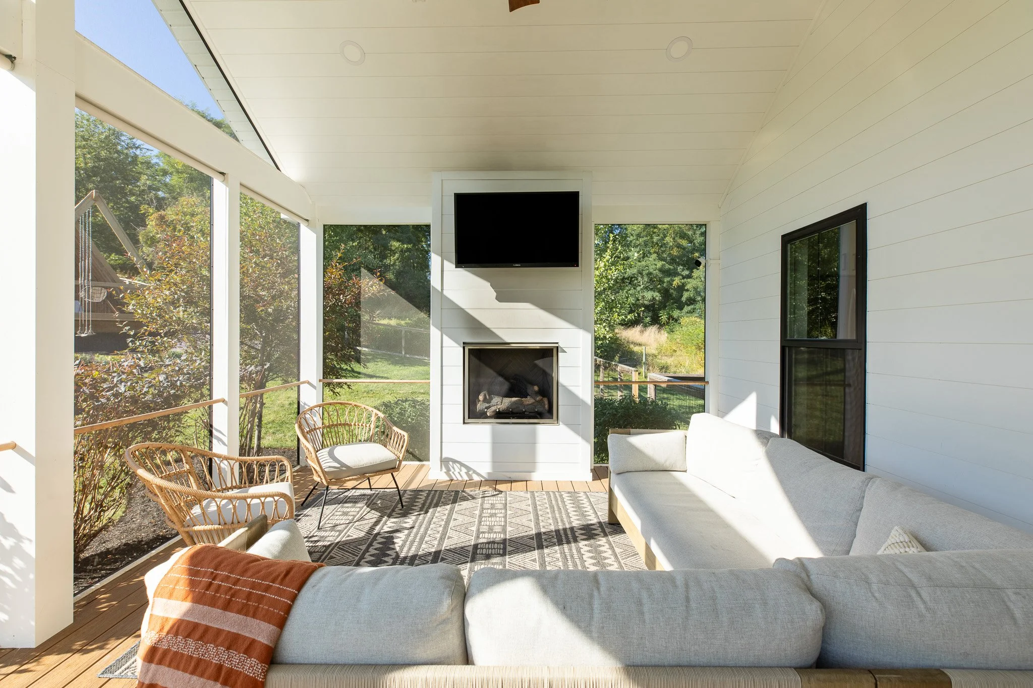 Sunlit screened porch with white sofa, two rattan chairs, a patterned rug, wall-mounted TV above a fireplace, and windows showing outdoor greenery.