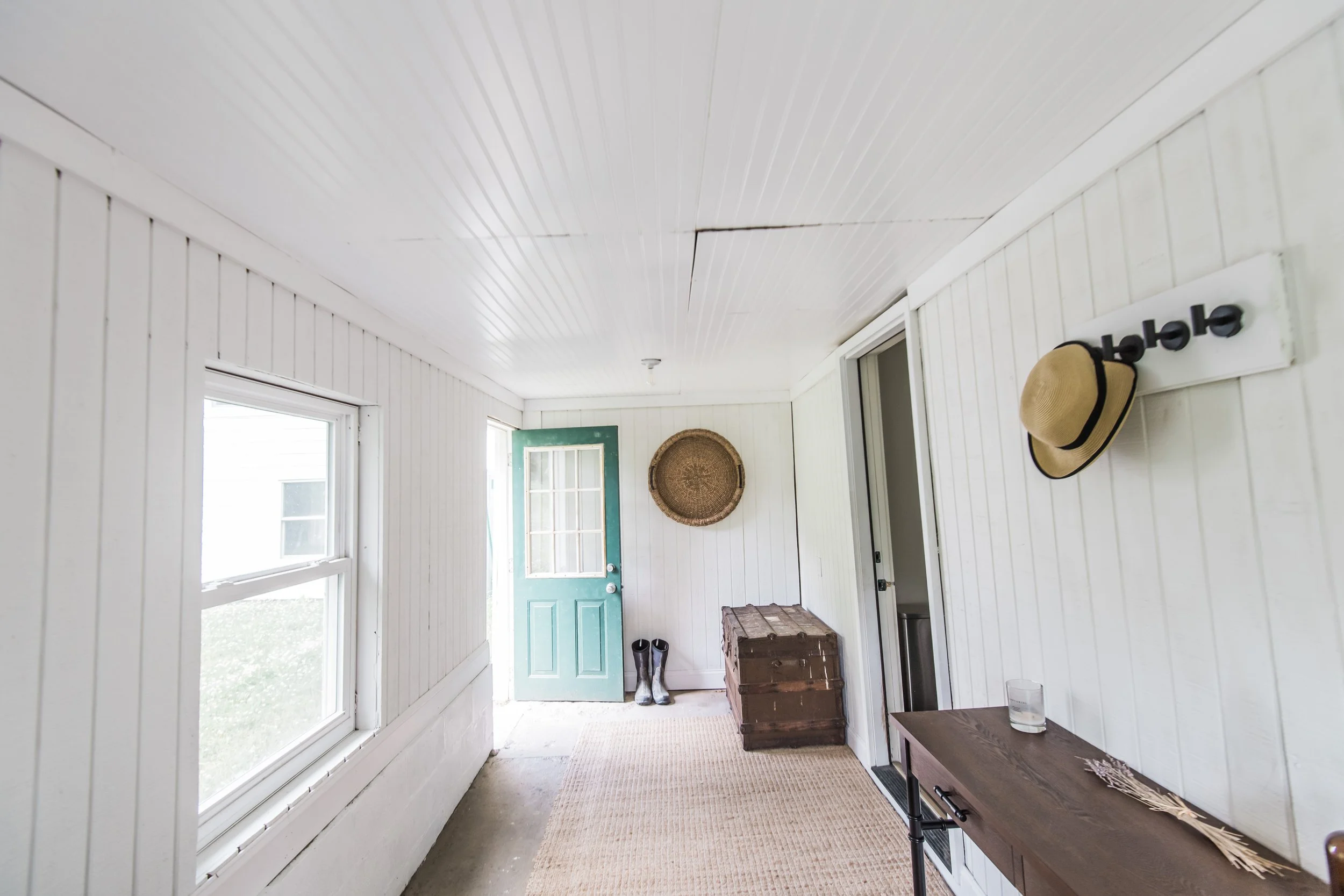 Bright porch with white wooden paneled walls, two windows, a teal door, a woven wall hanging, a chest, rain boots, and a wooden table with a glass of water and decorative sticks.