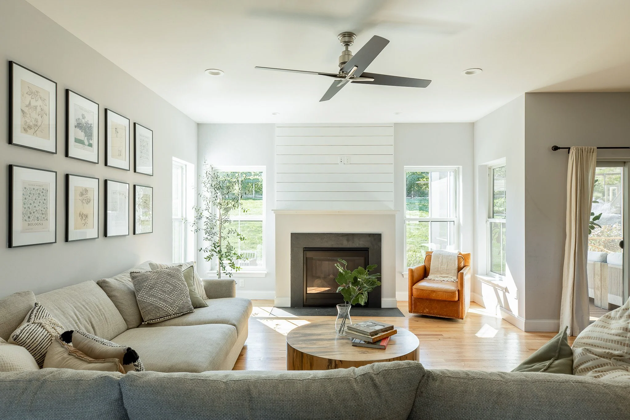 Bright living room with white walls, a fireplace, a ceiling fan, large windows, a beige couch with pillows, a round wooden coffee table with books and a plant, and an orange armchair near the window.