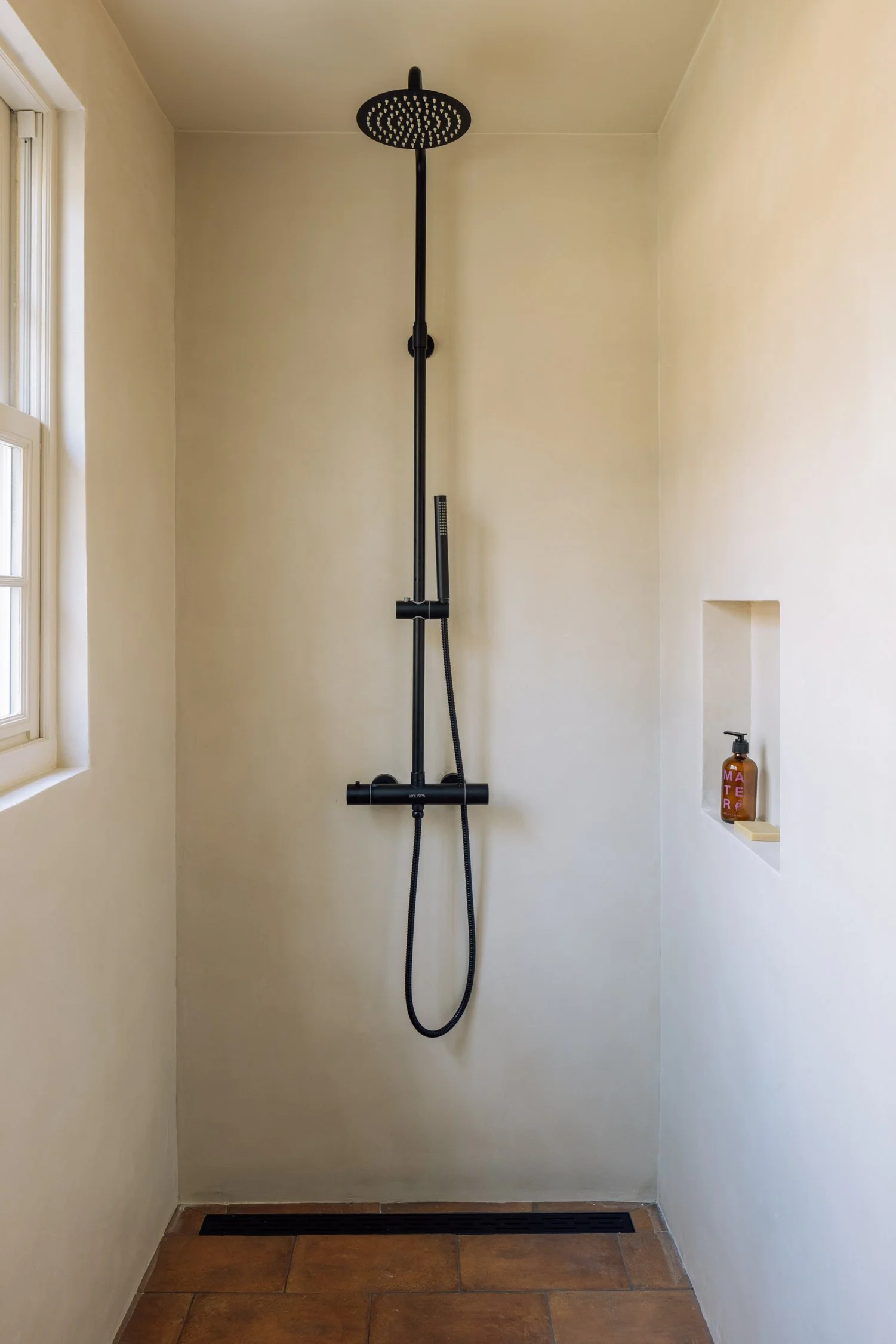 A modern shower with a black rain showerhead, a black handheld shower, and a niche with a soap dispenser in a beige tiled bathroom.