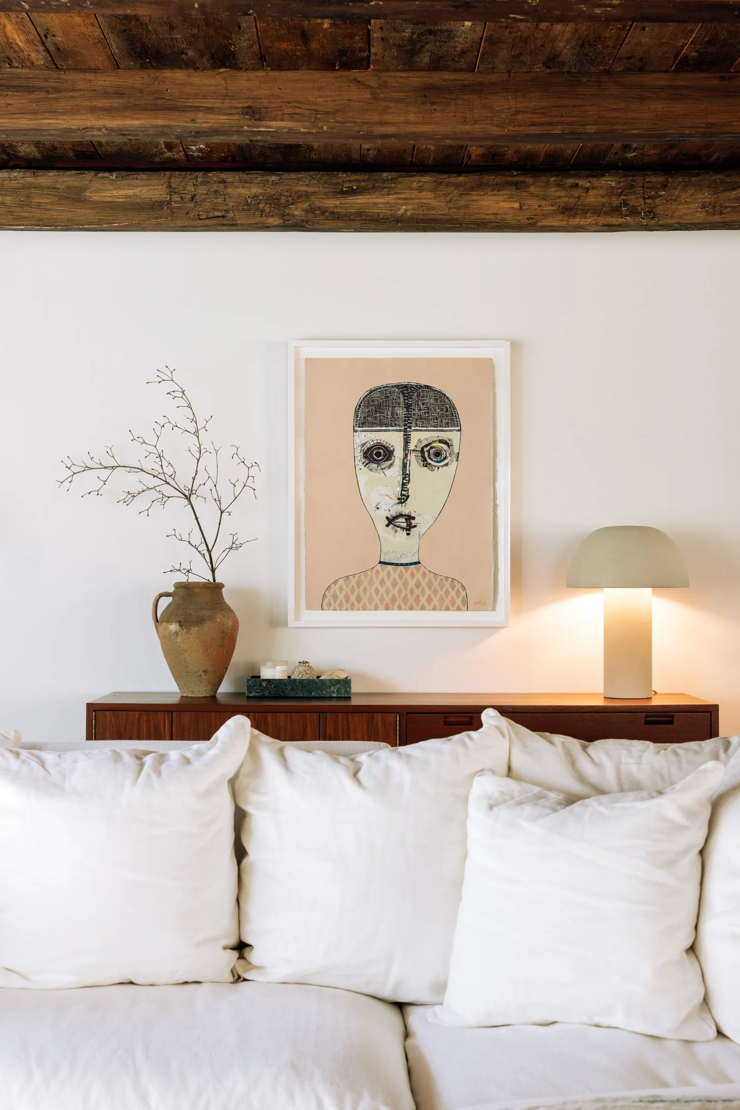 A cozy living room with a white couch, a wooden sideboard, a modern beige table lamp, a framed abstract portrait art piece, a decorative vase with a branch, and a tray with small items on top.