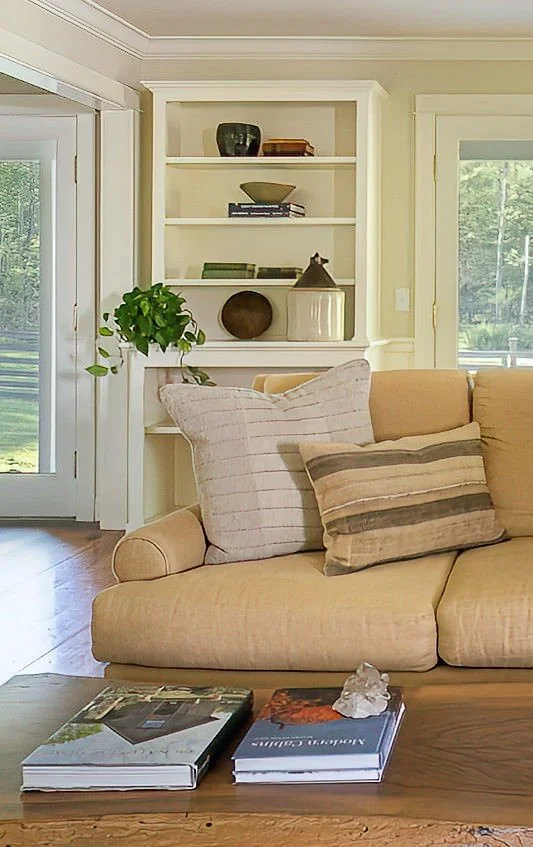 Living room with a beige couch, striped pillows, a wooden coffee table with magazines and a crystal, and a white bookshelf with decorative items and a green plant nearby.