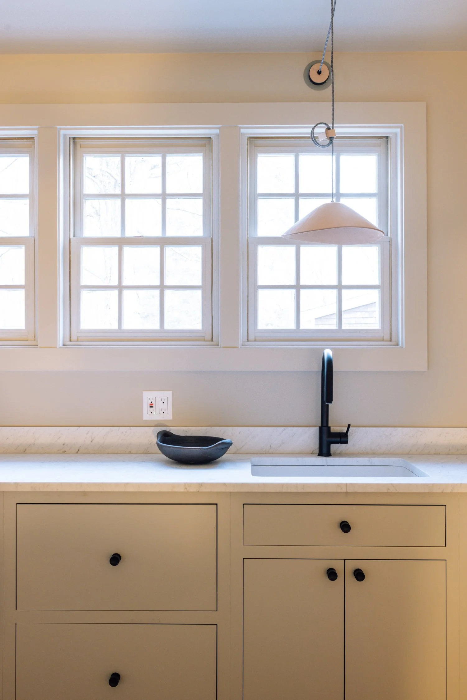 Kitchen with three windows, beige cabinets, a black sink fixture, a marble countertop, and a beige pendant light hanging from the ceiling.