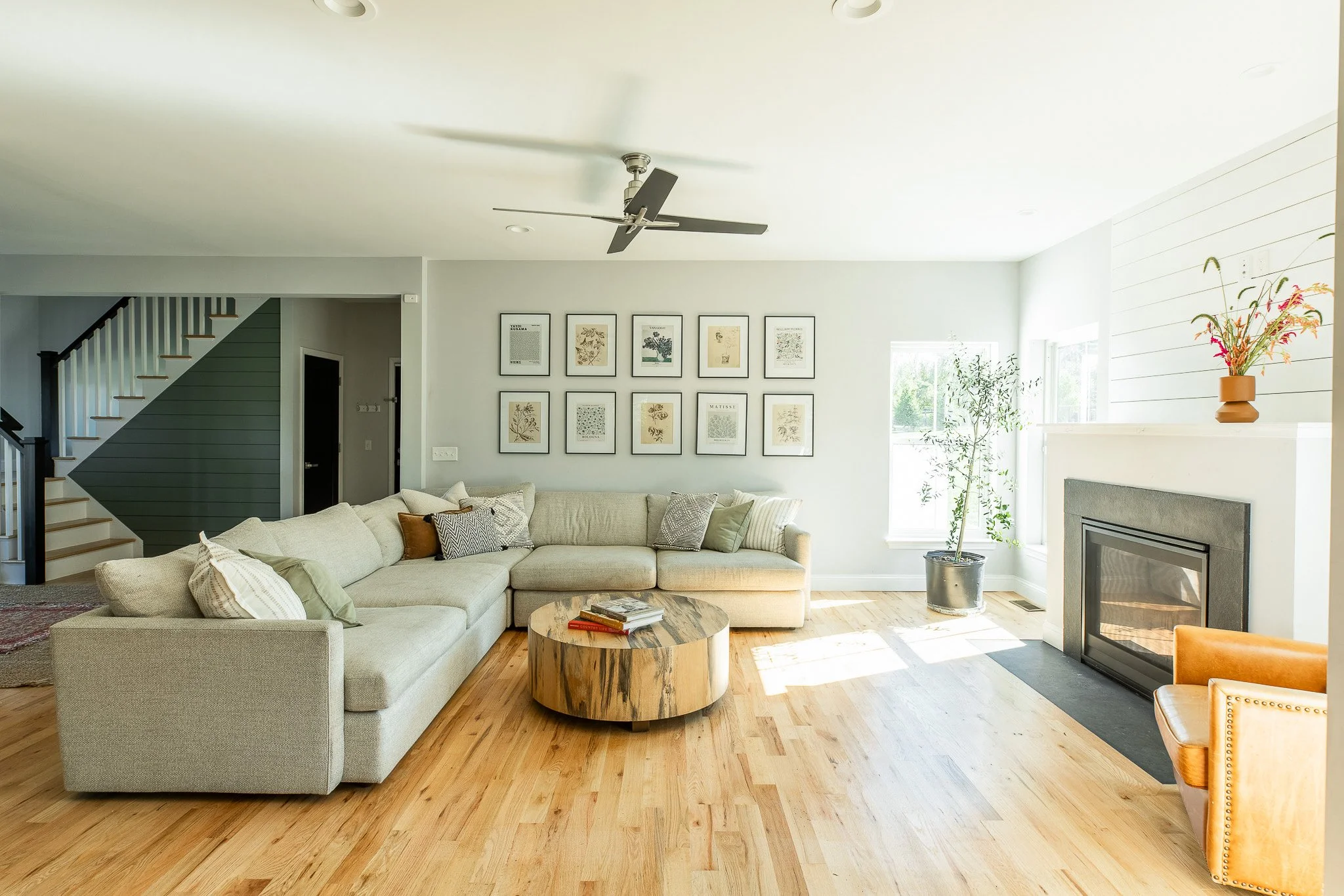 Living room with a large beige sectional sofa, wooden coffee table, fireplace, and framed artwork on the wall. Bright natural light coming through windows, with a potted plant near the window and a vase with flowers on the mantle.