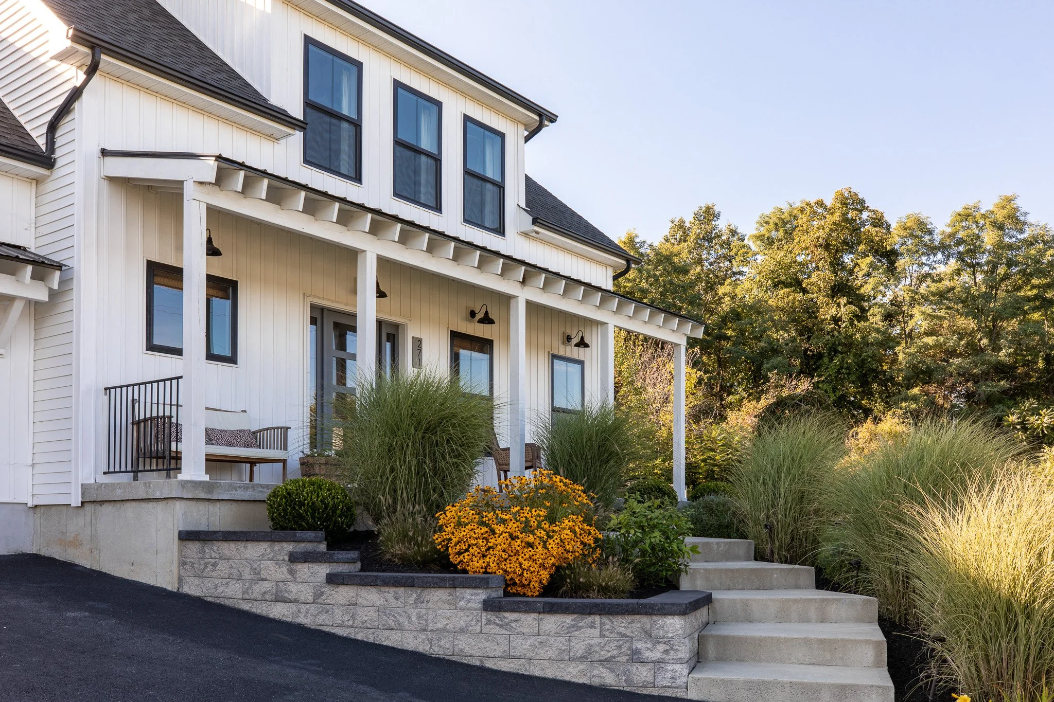 A white modern house with a small front porch, landscaped with yellow flowers, green shrubs, and tall grass, with a sidewalk and trees in the background.