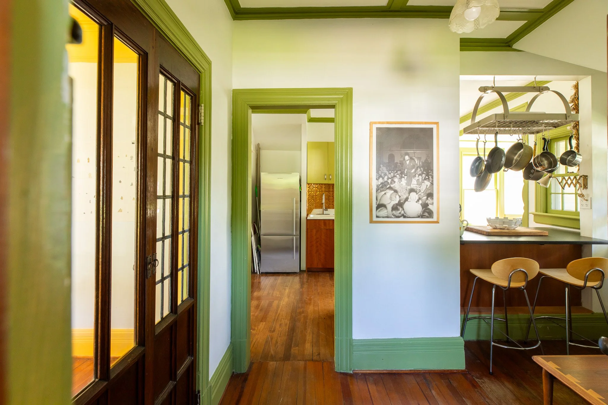 Interior view of a house with kitchen and dining area, green trim, wooden floors, and black-and-white photograph on wall.