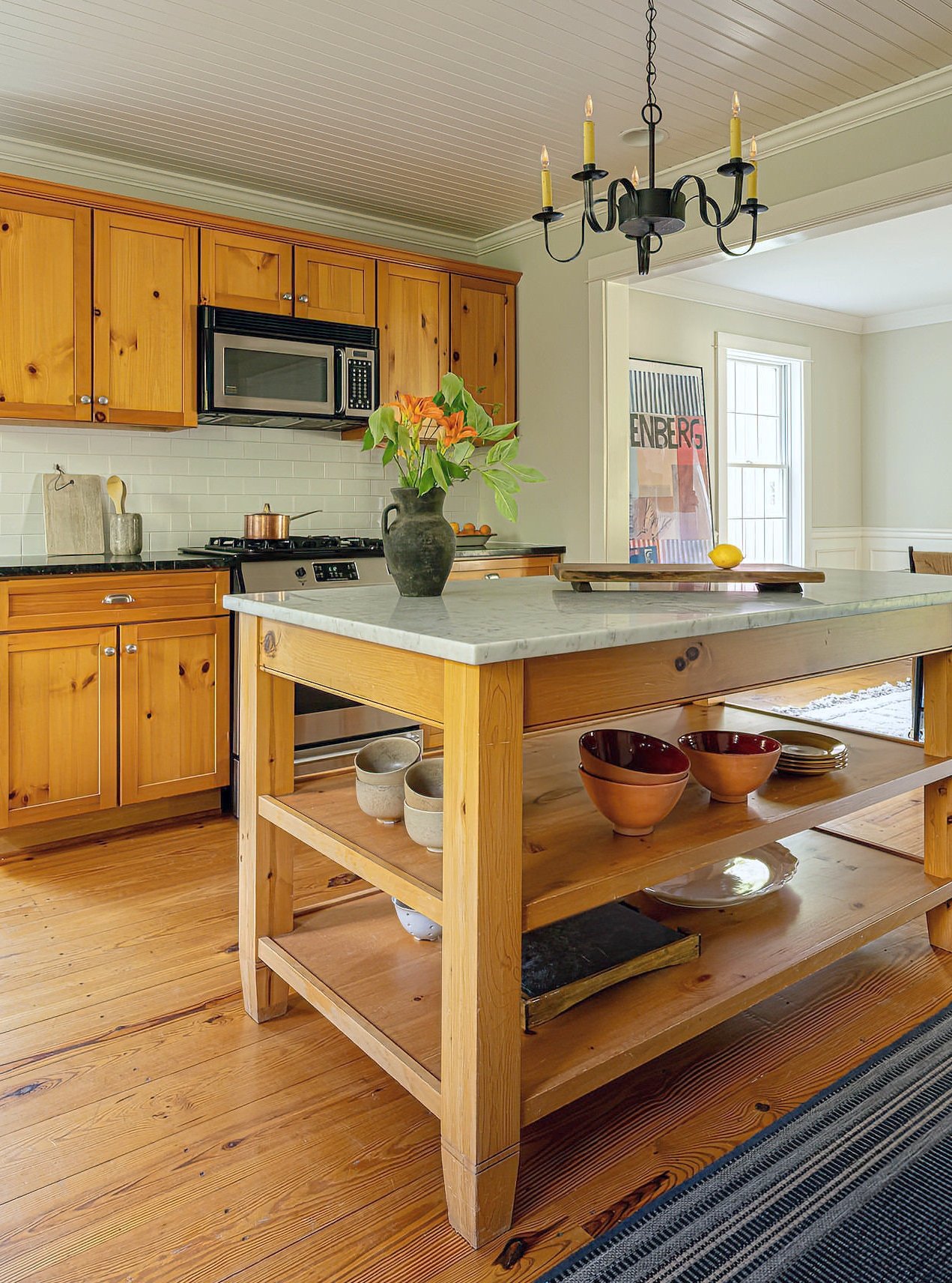 Kitchen with wooden cabinets, a marble-topped island, a chandelier, and a window with a framed poster leaning against the wall.