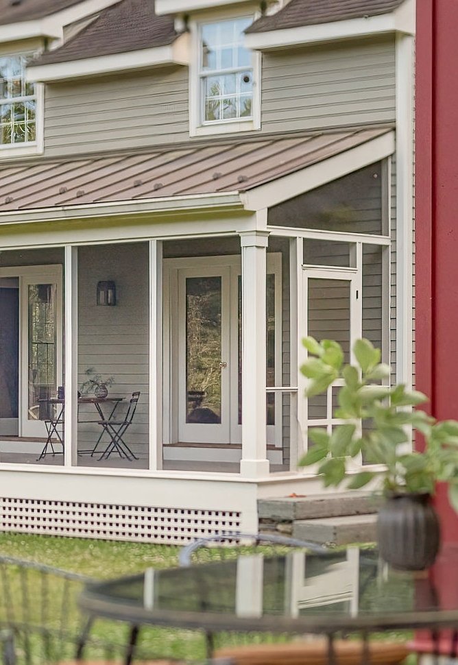 A screened porch attached to a house with gray siding and white trim, with an outdoor patio table and chairs visible inside, and a potted plant on a table in the foreground.