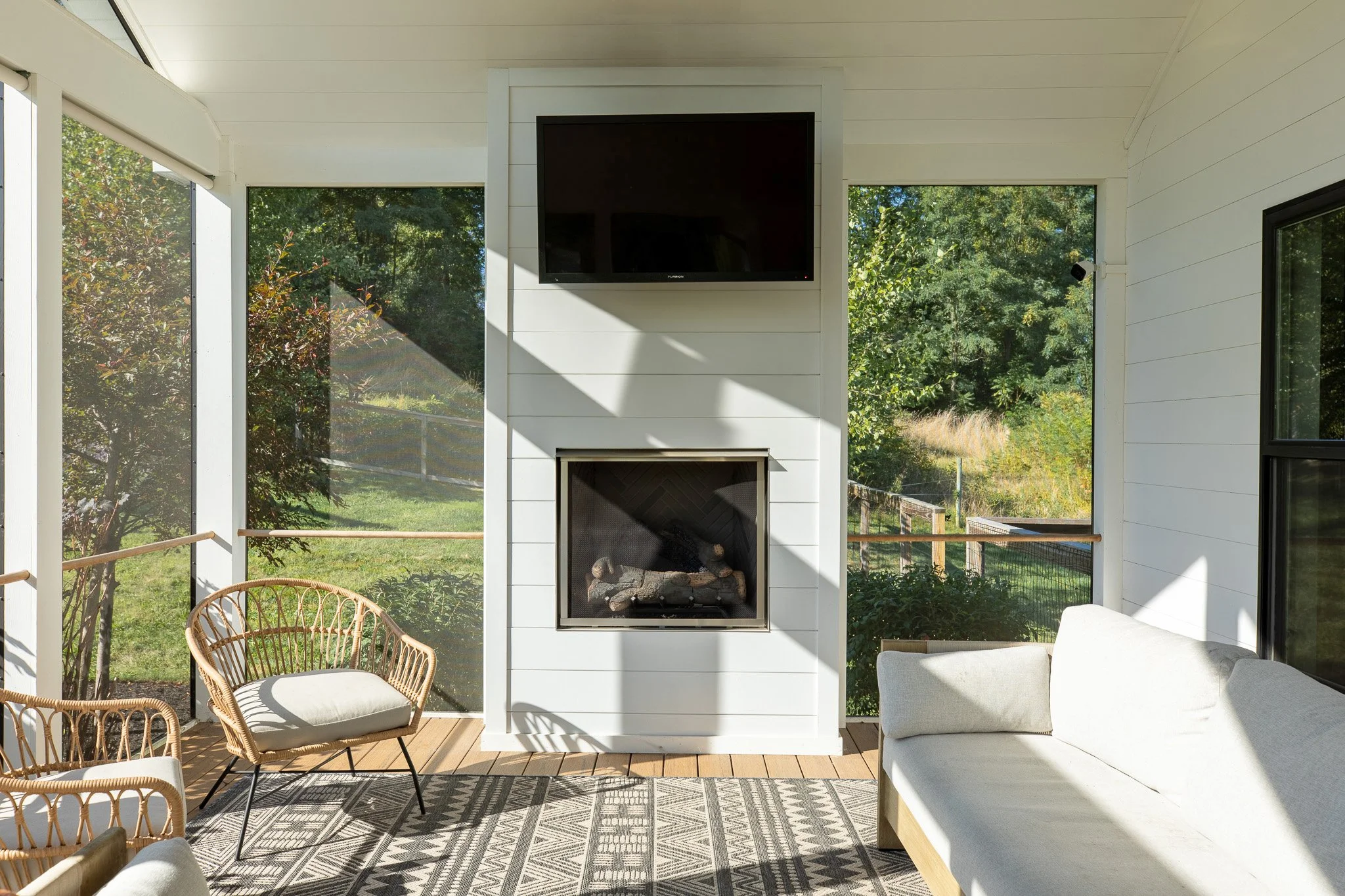Sunlit screened porch with white walls, a mounted TV above a fireplace with logs, a white sofa, and rattan chairs on a patterned rug, overlooking a green backyard.