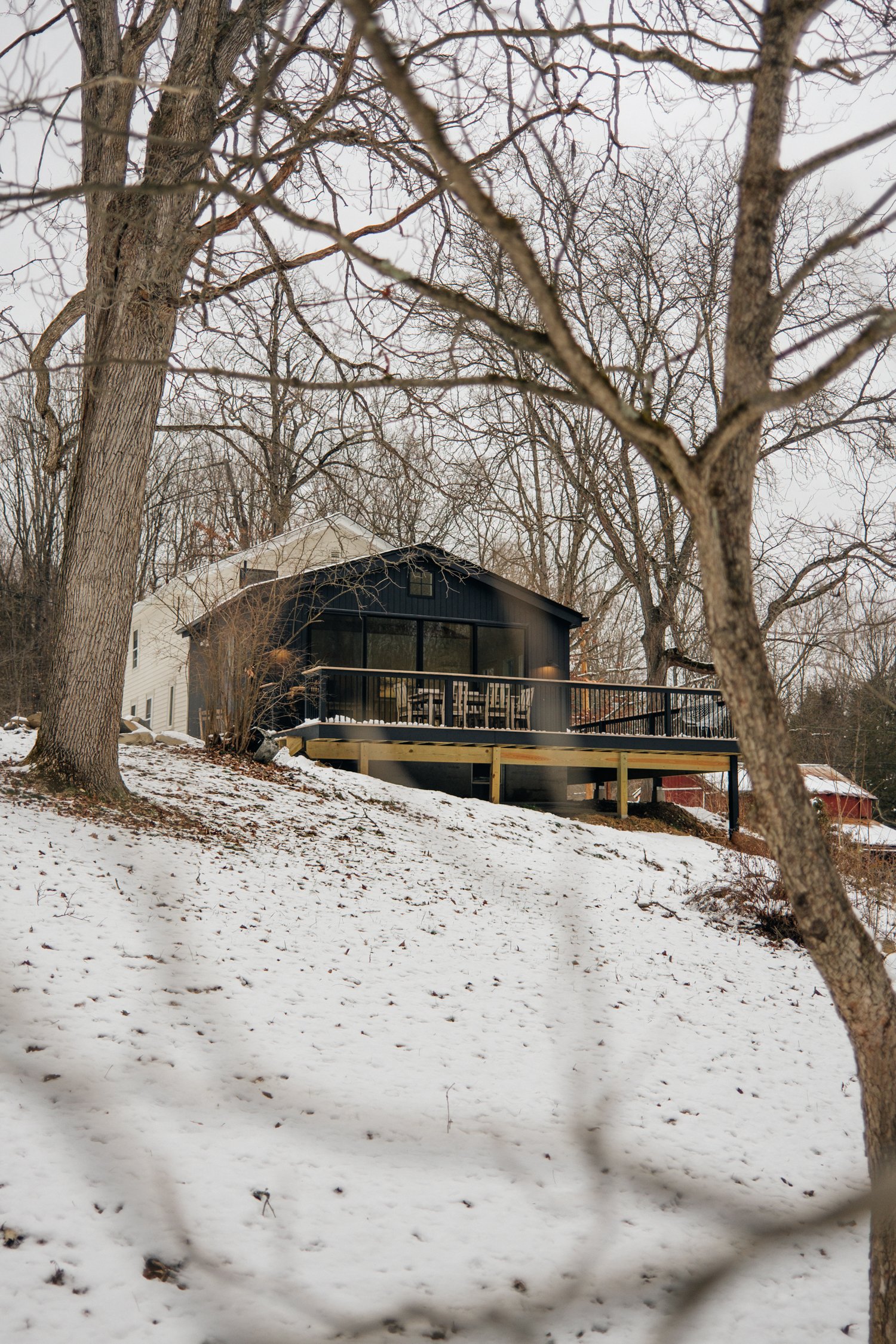 A house with large windows and a wooden deck is set on a snow-covered hillside surrounded by leafless trees.
