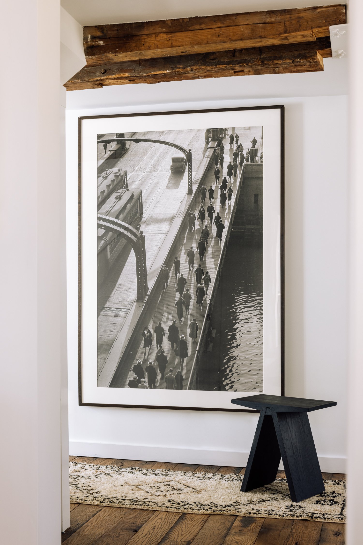 Black and white framed photograph of a busy pedestrian bridge over water, seen from above, with people walking and trucks on the adjacent road.