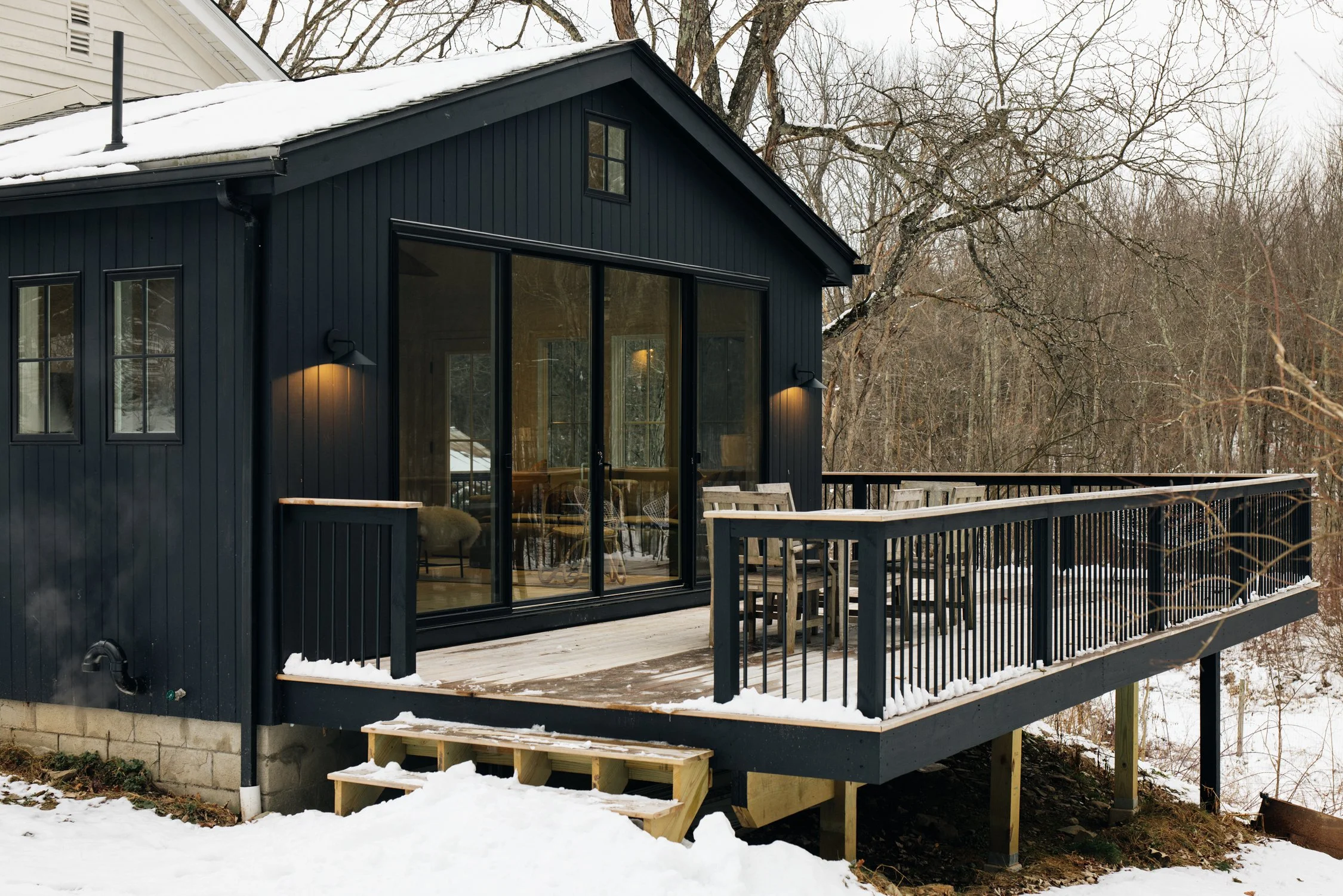 Black wooden house with large glass sliding doors and a snow-covered deck with outdoor furniture, surrounded by snow and leafless trees.