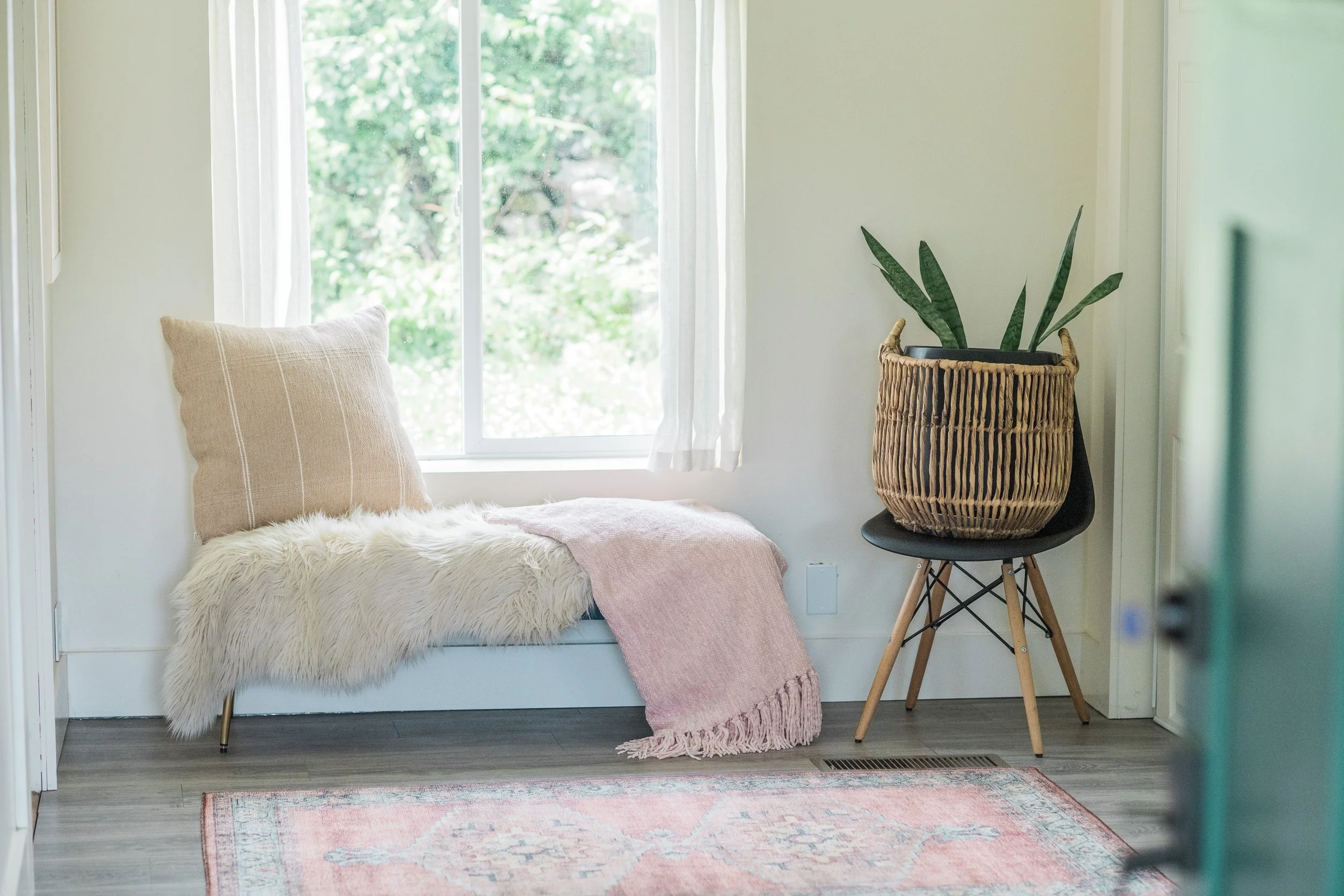 A cozy corner with a window seat adorned with pillows and a throw blanket, a black chair with a basket containing a plant on top, and a pink patterned rug on the wooden floor.