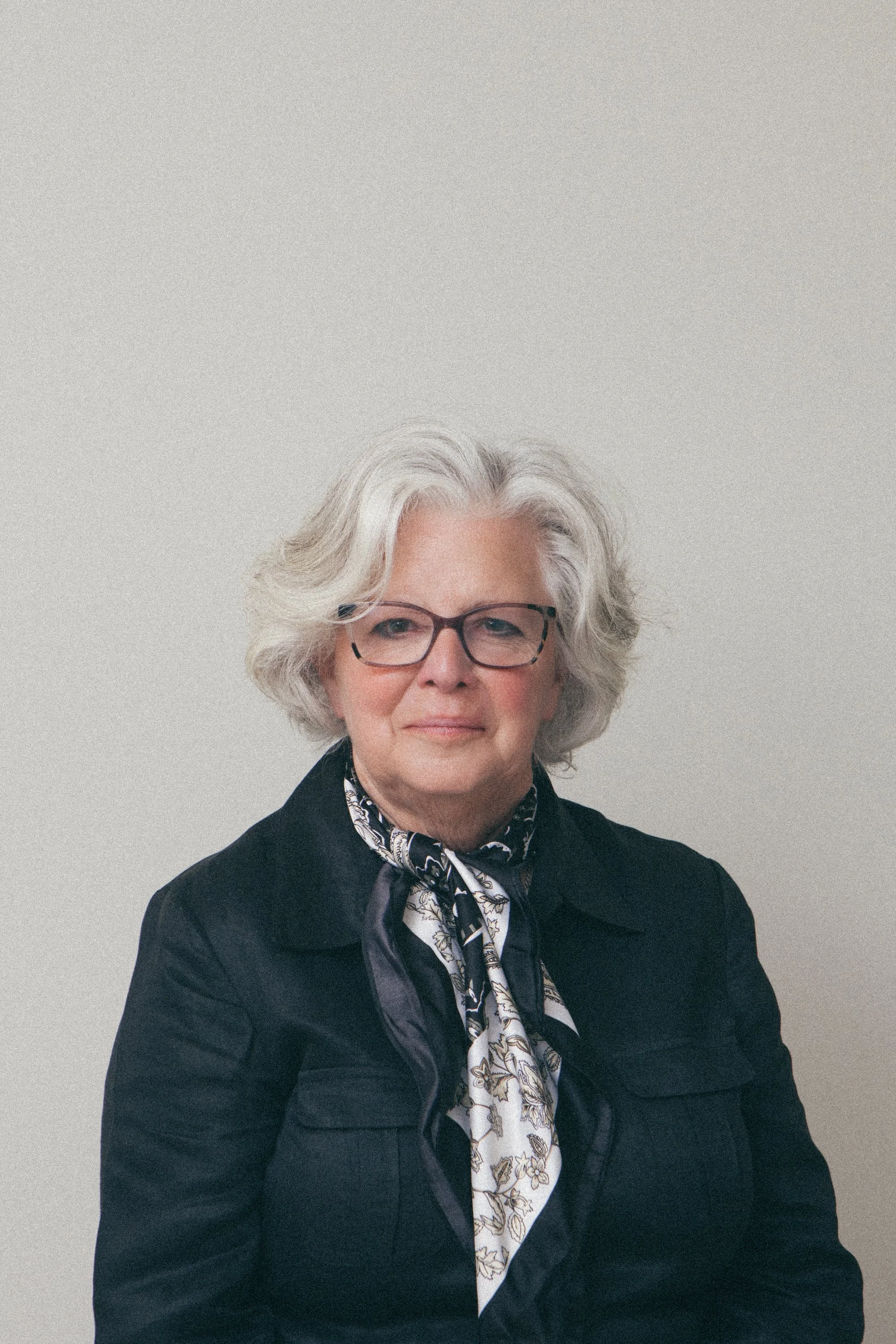 An older woman with short gray hair and black glasses dressed in a gray top with black beaded necklace, standing outdoors with trees and a building in the background.