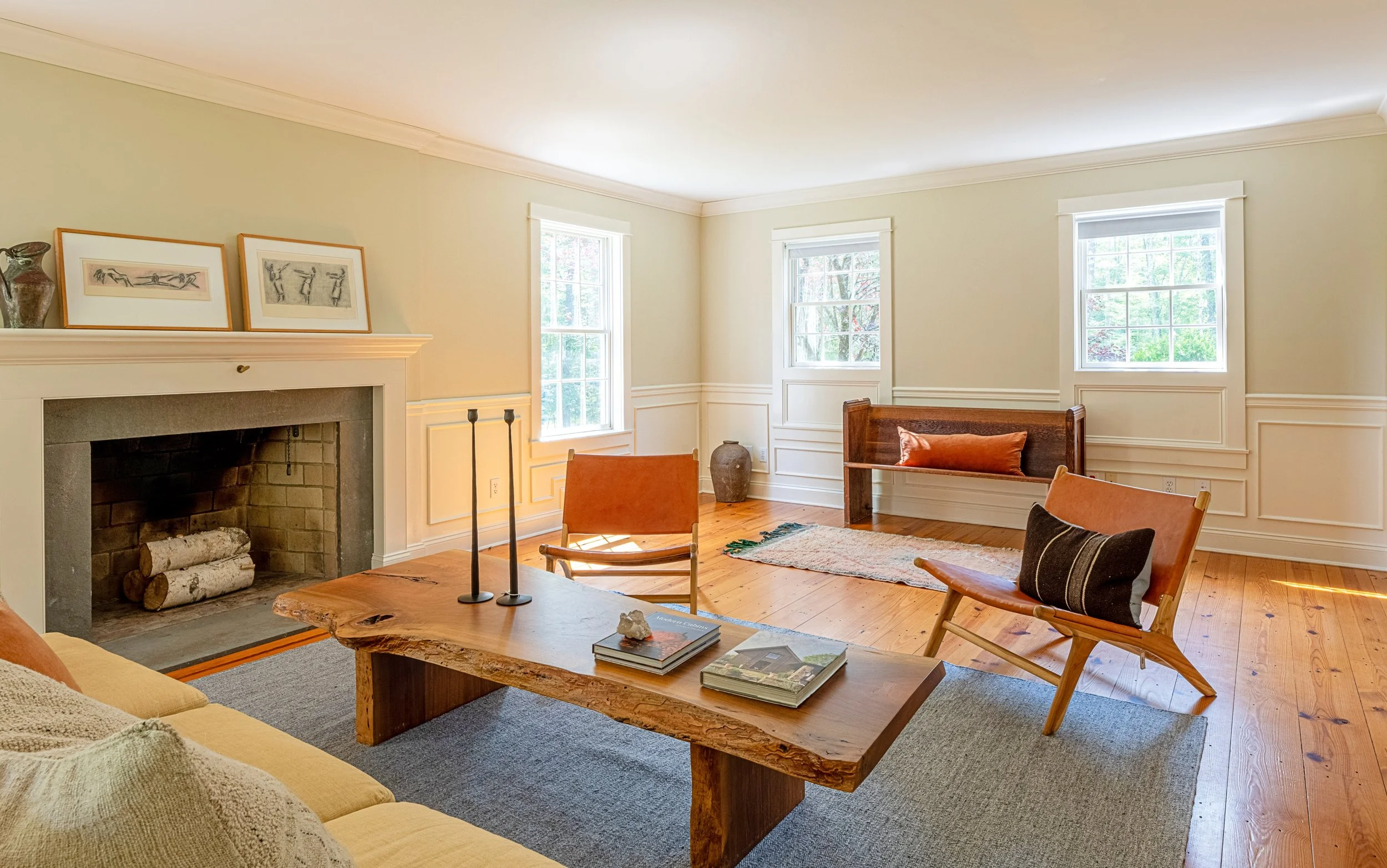 Living room with cream walls, three windows, a fireplace with logs, a wooden coffee table, a yellow sofa, and various chairs with cushions.
