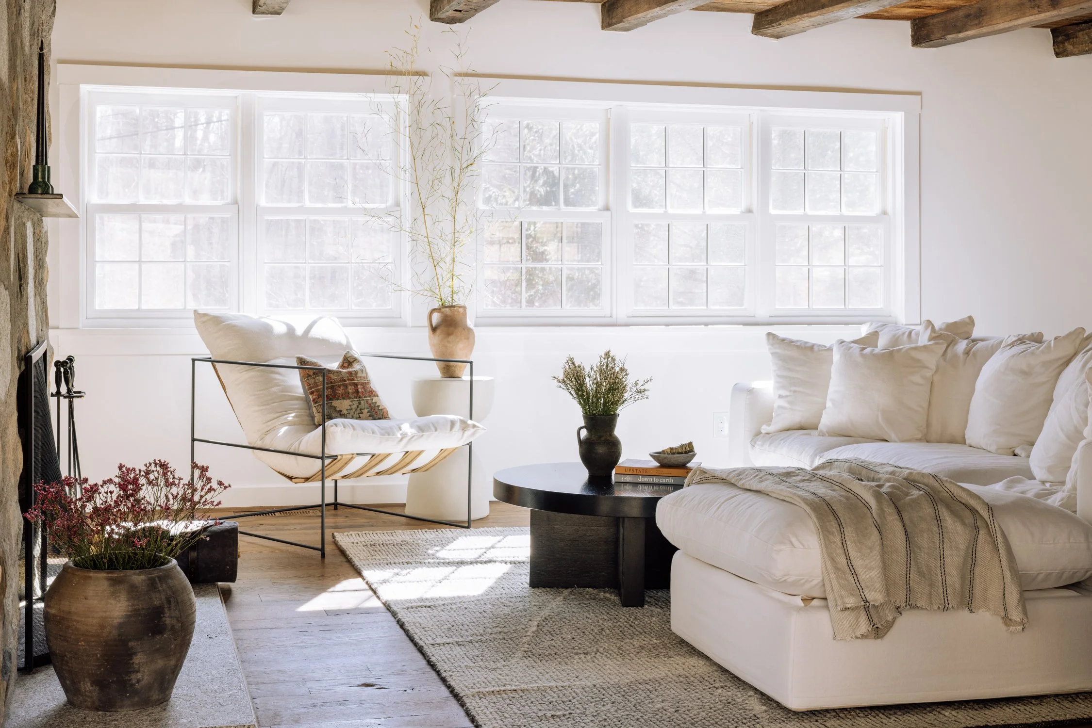Bright living room with white sofa, round black coffee table, armchair with pillow, large window with sunlight, stone fireplace, potted plants, and wooden ceiling beams.