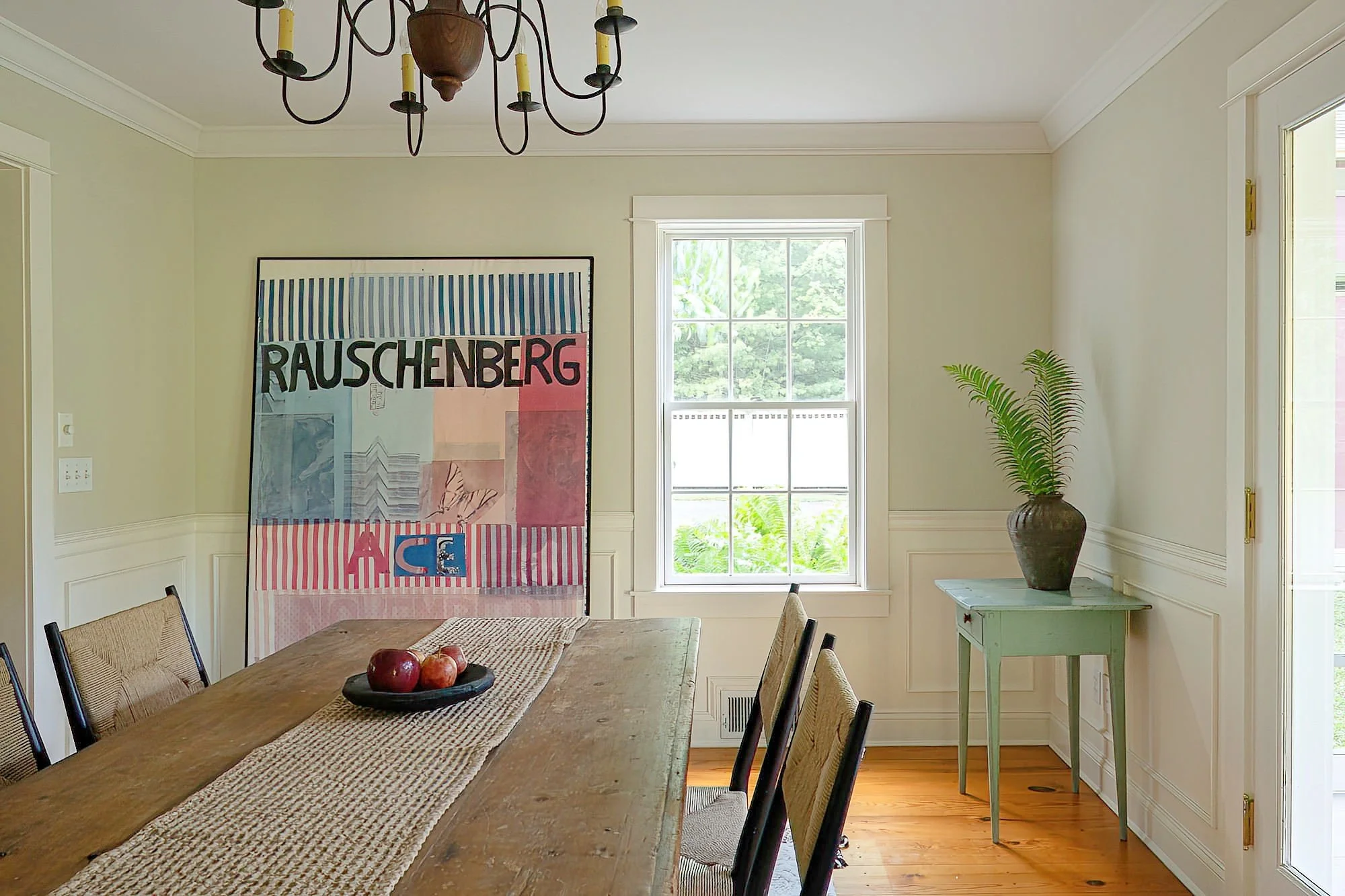 A room with a wooden dining table, six chairs, a large pink and blue artwork on the wall, a window with green outside view, a green side table, and a potted plant in a brown vase.