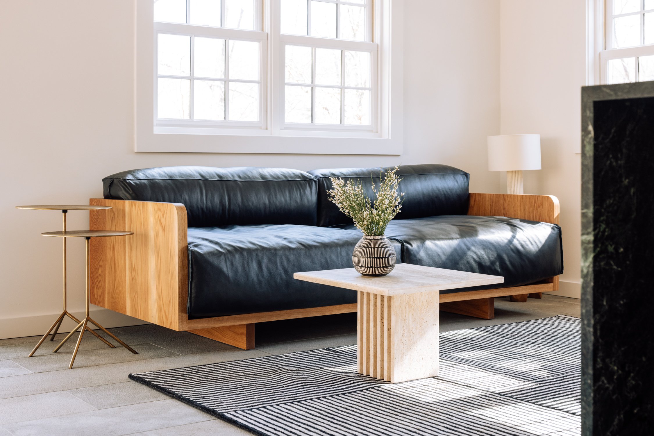 Living room with a black leather sofa, wooden accents, a small marble side table, and a industrial style striped rug, beside a window with natural daylight.