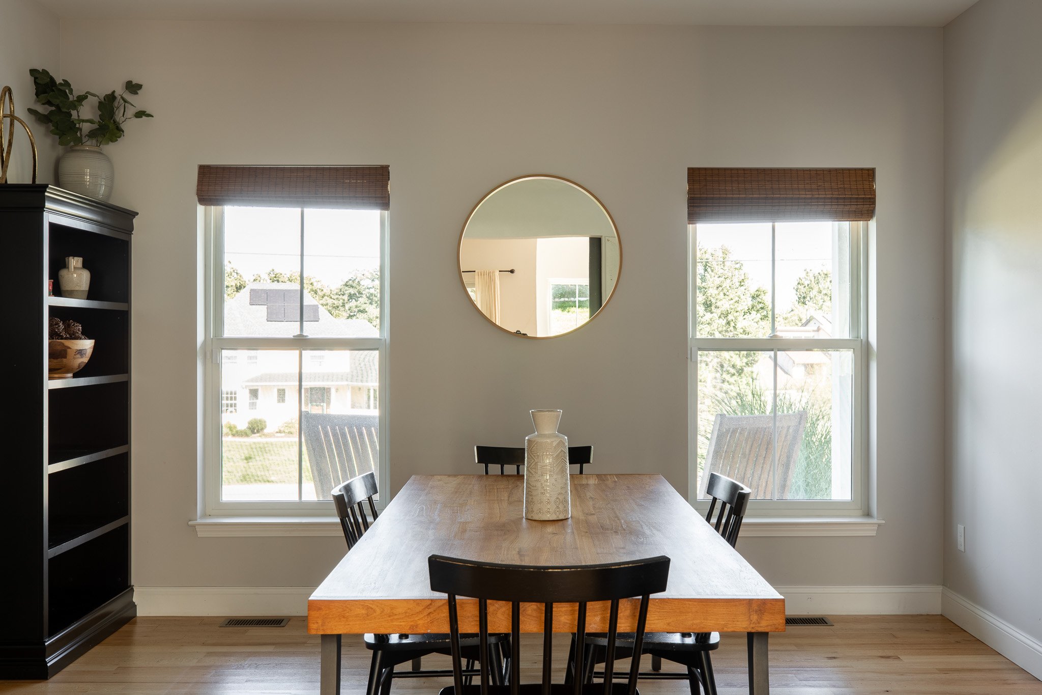 Dining room with wooden table, black chairs, a round mirror on the wall, two windows, and a black bookshelf with decorative items.