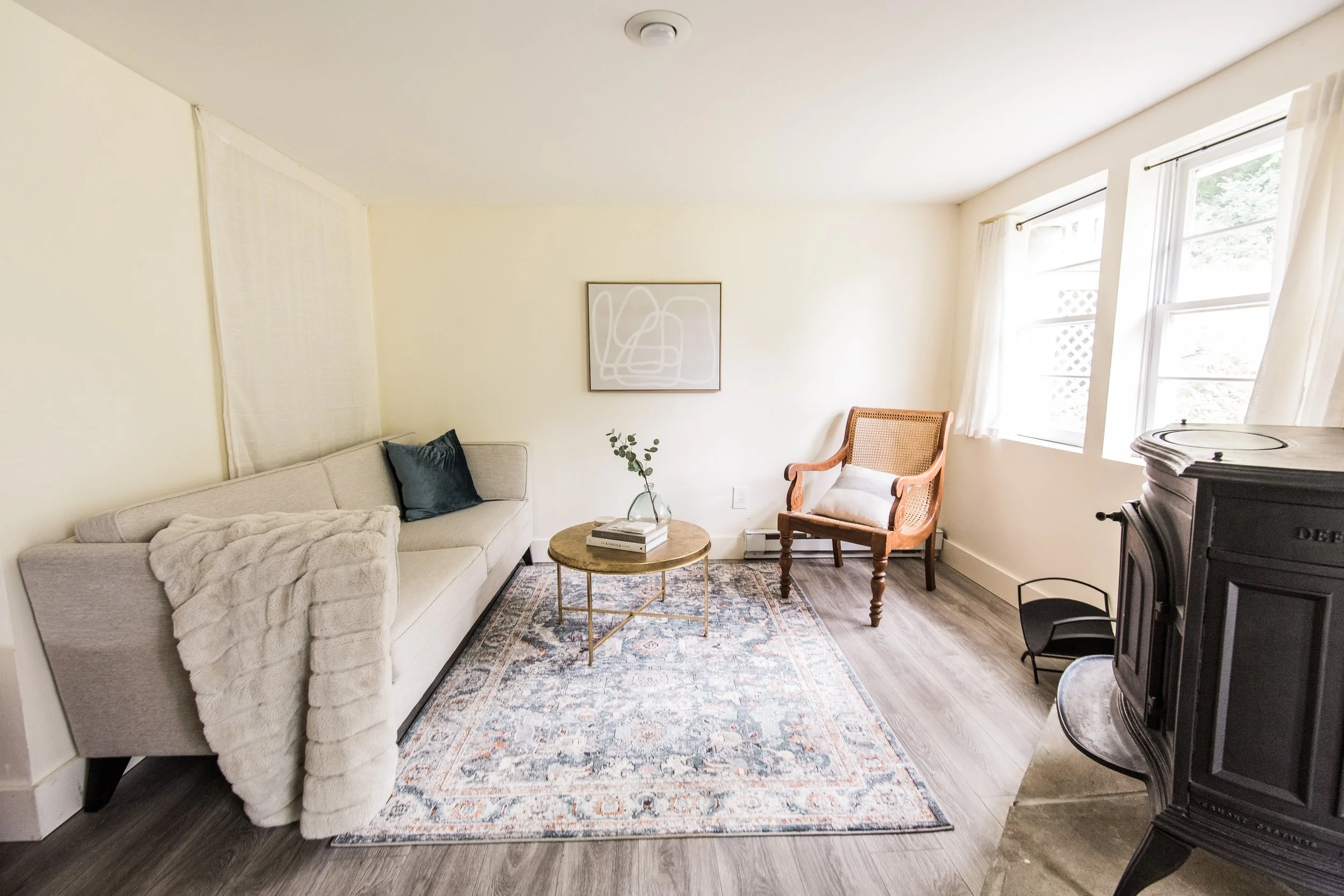 Living room with a beige sofa, coffee table, patterned rug, armchair, and a wood stove, in front of cream-colored walls and windows with white curtains.