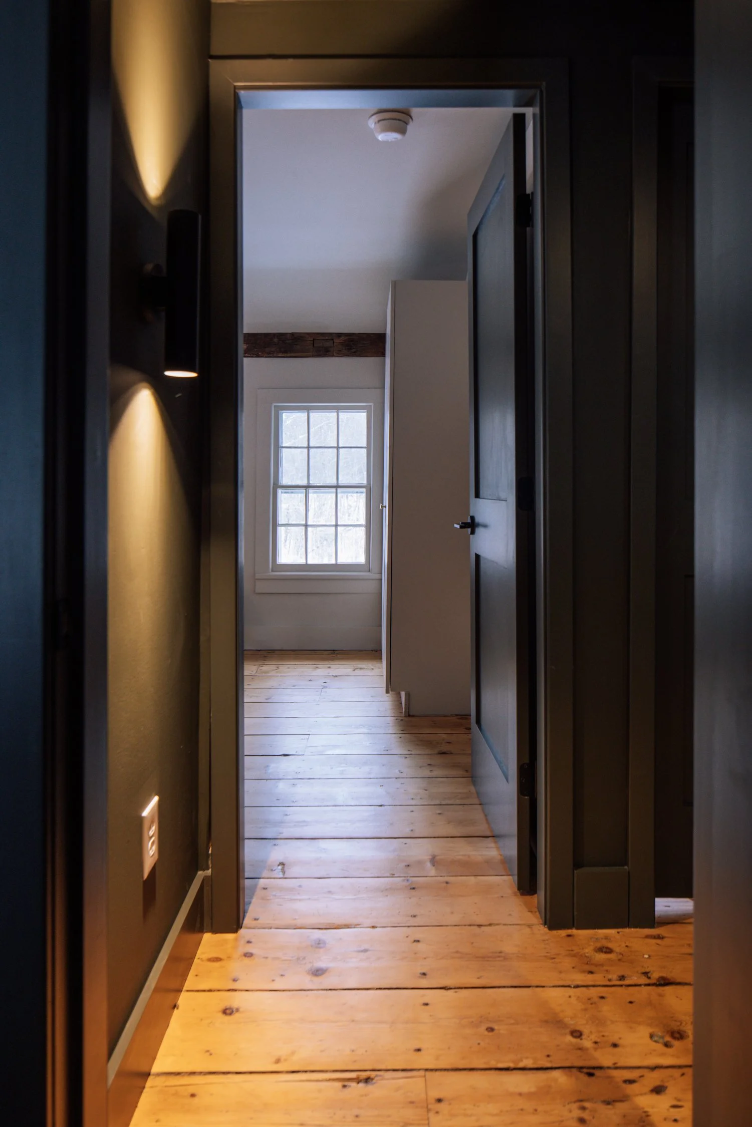 View through a doorway into a room with a window and wooden floorboards, with black walls and trim surrounding the doorway.