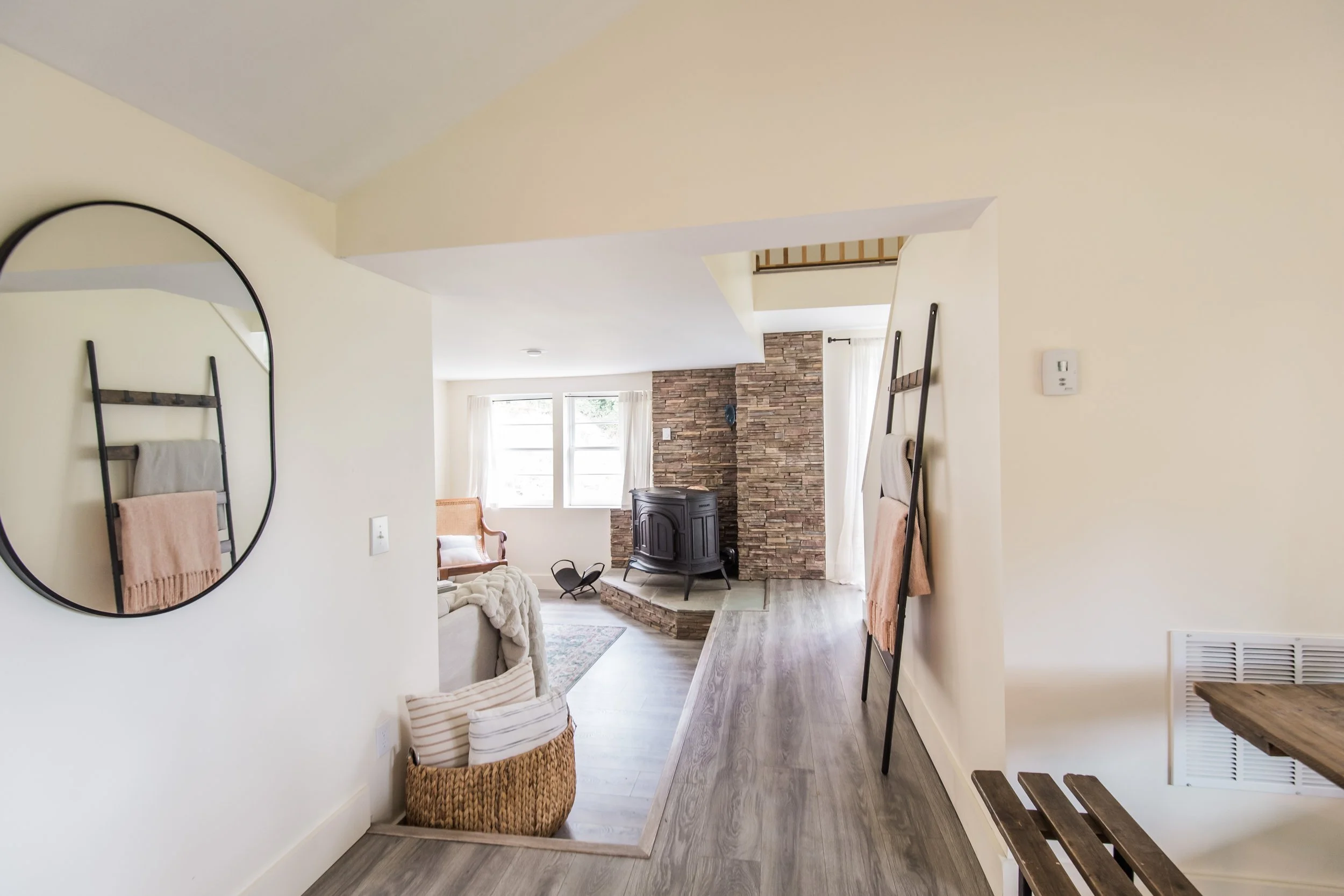 Living room with a stone fireplace, wooden flooring, and beige walls. A cozy armchair with white cushions, a round mirror on the wall, and a basket with pillows are visible. Large windows allow natural light into the room.