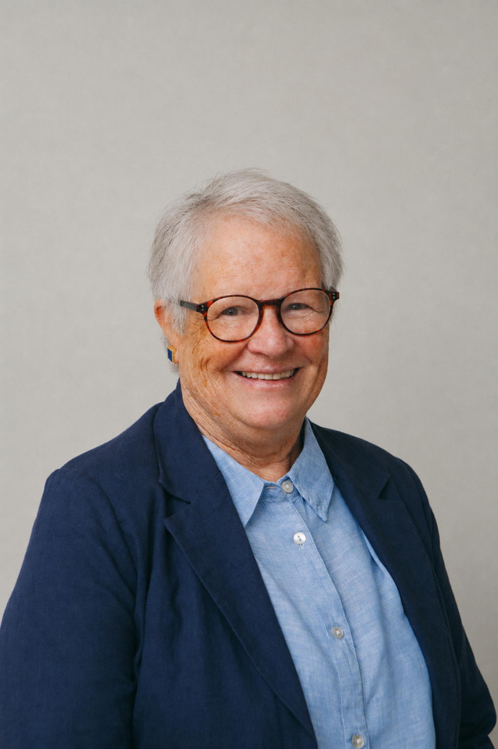 An elderly woman with short gray hair, glasses, and a black sweater with a turtleneck, holding a piece of paper and smiling, standing outdoors in a wooded area.