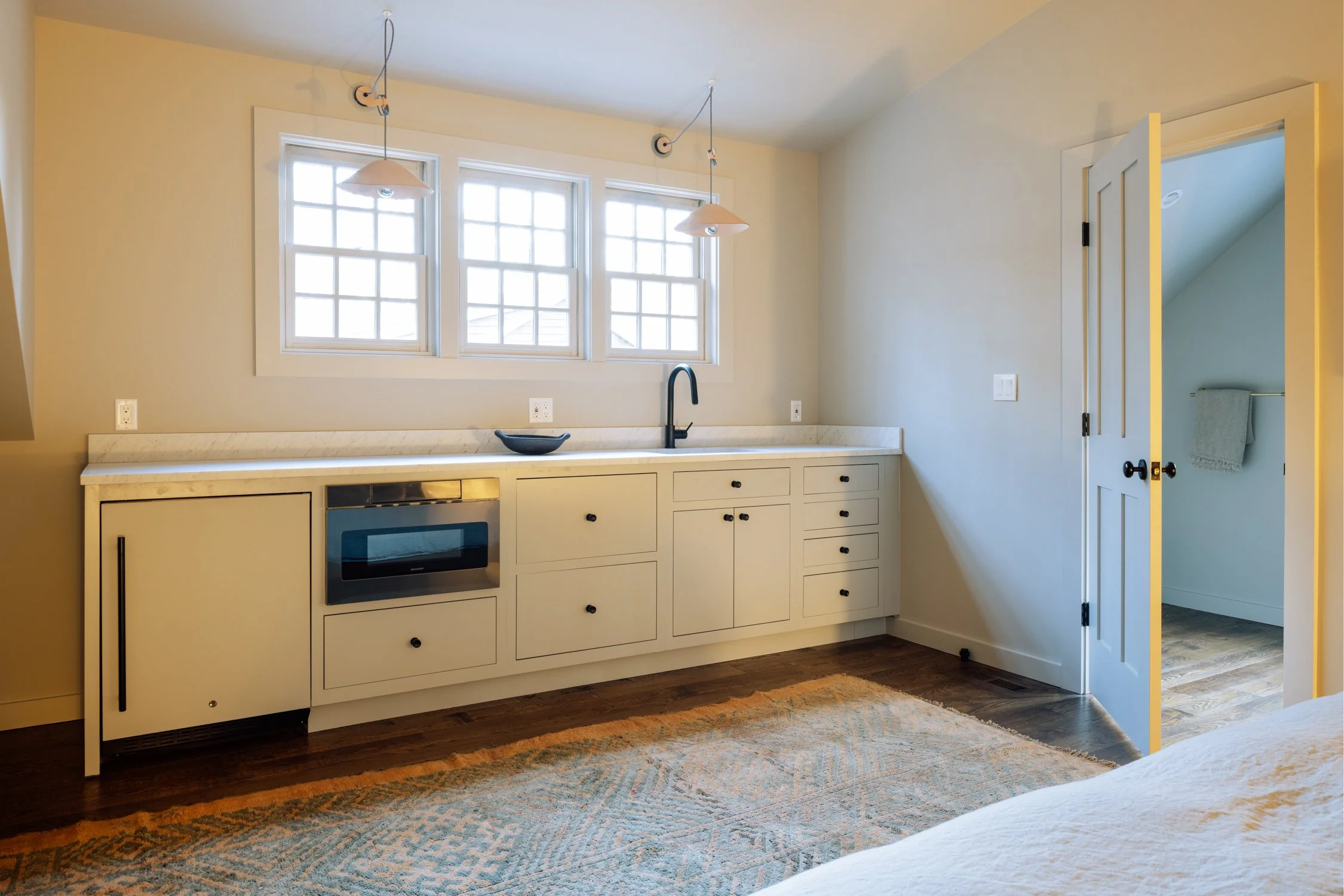 A small kitchenette with white cabinets, a marble countertop, and a black sink, illuminated by natural light from three windows. There is a closed door to a small room with a towel hanging on a rail.