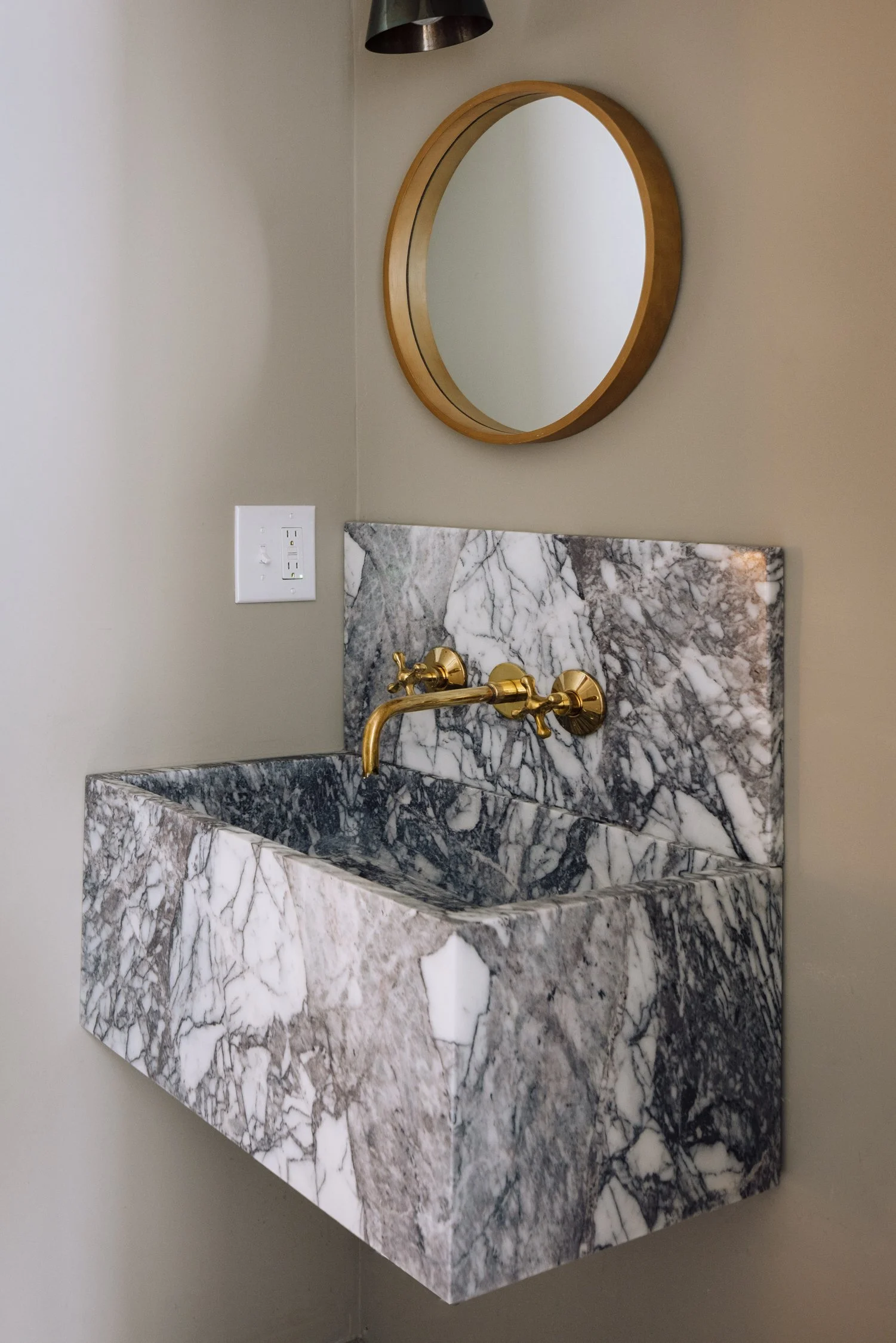 A wall-mounted marble sink with golden faucet, a round wooden mirror above it, and a black wall-mounted light fixture. There is also a power outlet on the beige wall.
