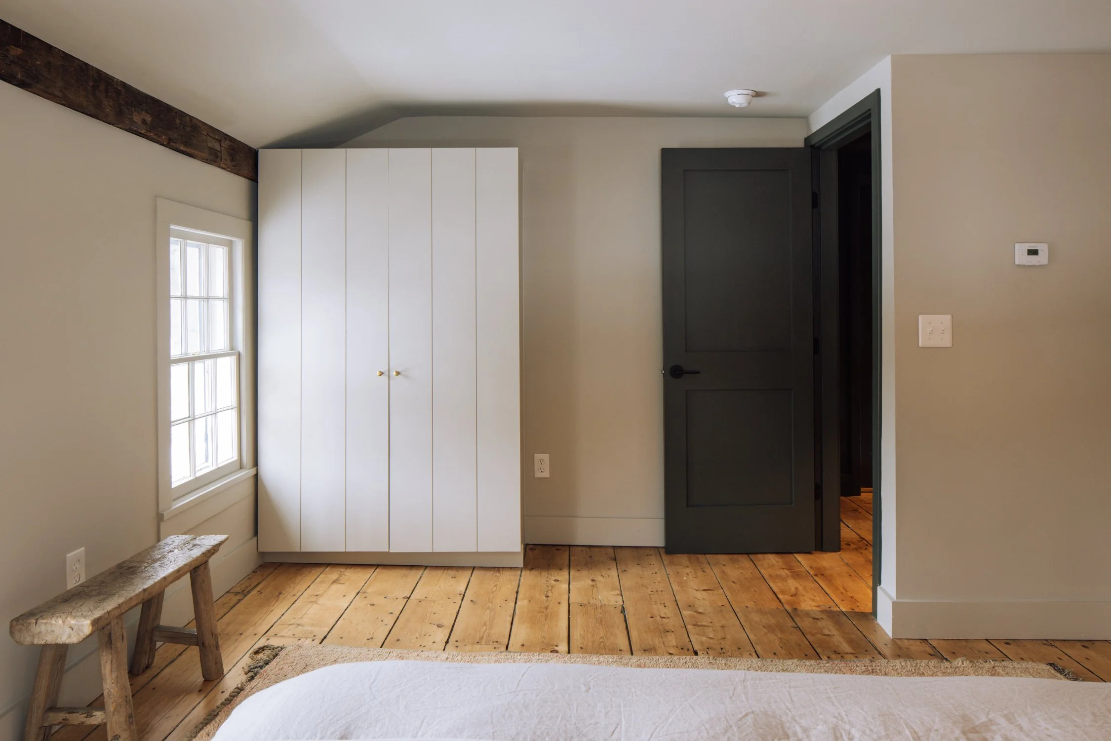Interior view of a bedroom corner with a window, white closet doors, a wooden bench, beige walls, and a dark gray door on a wooden floor.
