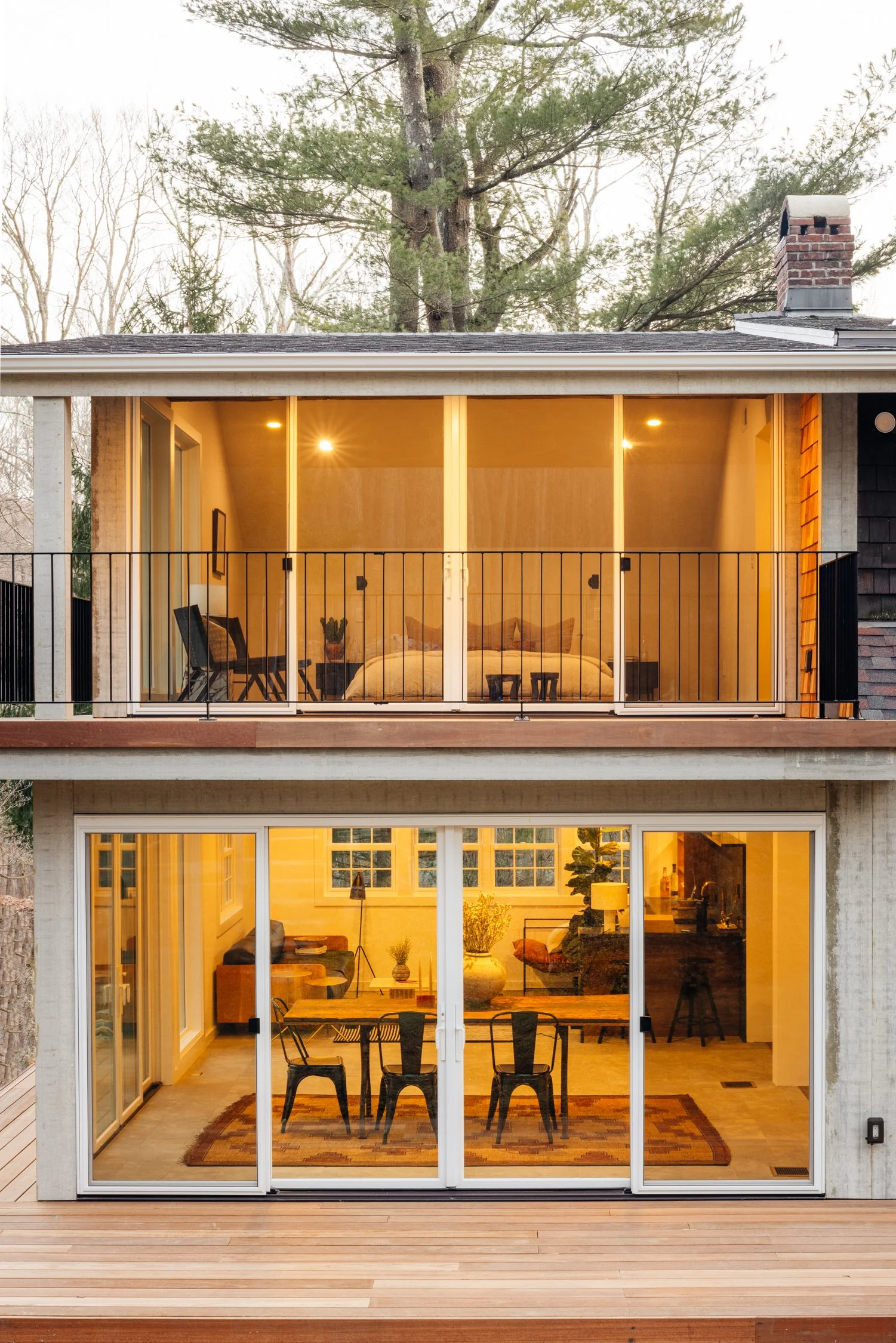 A two-story modern house with a large glass door and windows, a balcony with a railing on the upper level, and a wooden deck outside. Inside, warm lighting illuminates a bedroom upstairs and a dining area downstairs.