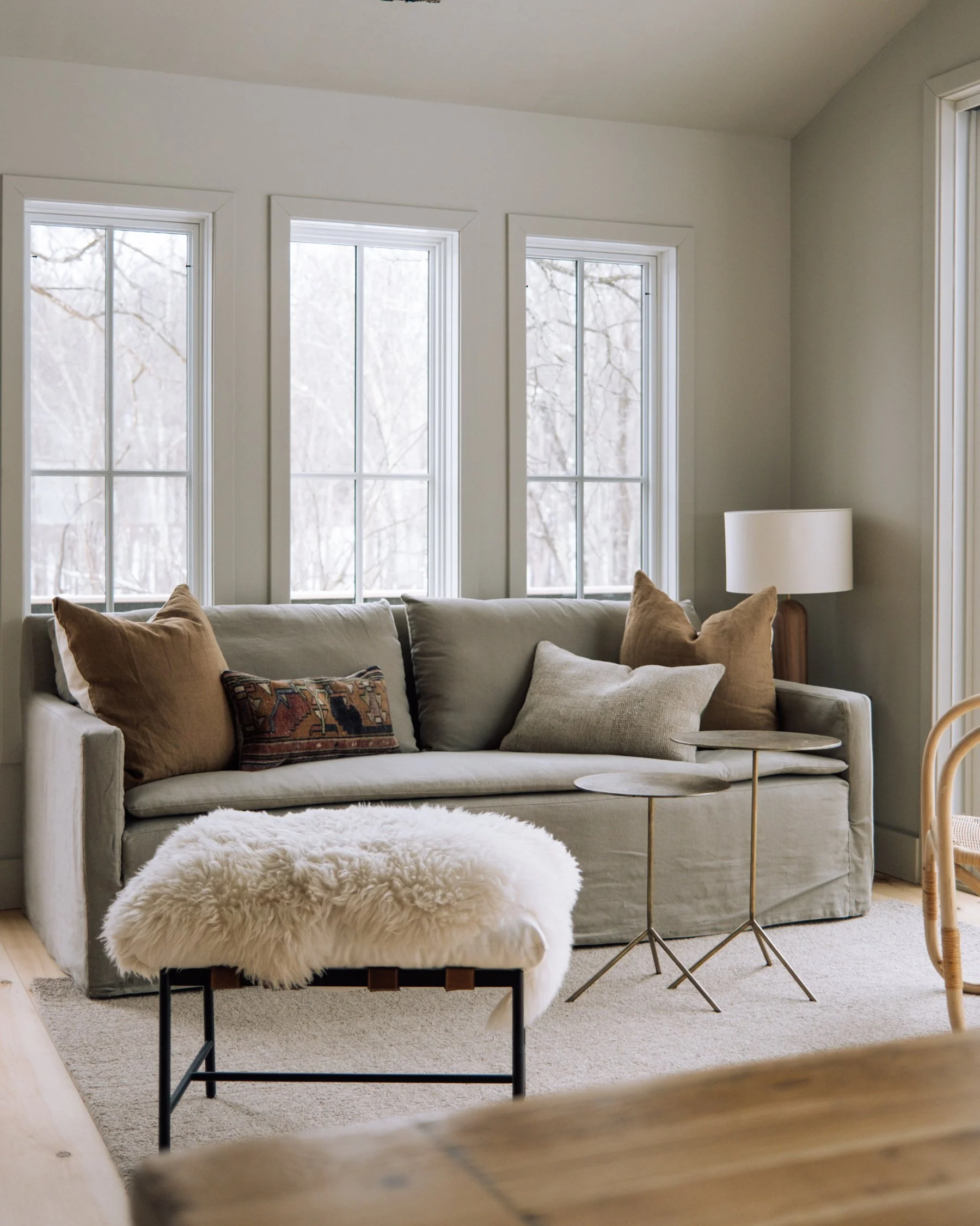 Living room with beige sofa, multiple pillows, sheepskin ottoman, side tables, and a lamp near three large windows showing a snowy outdoor scene.