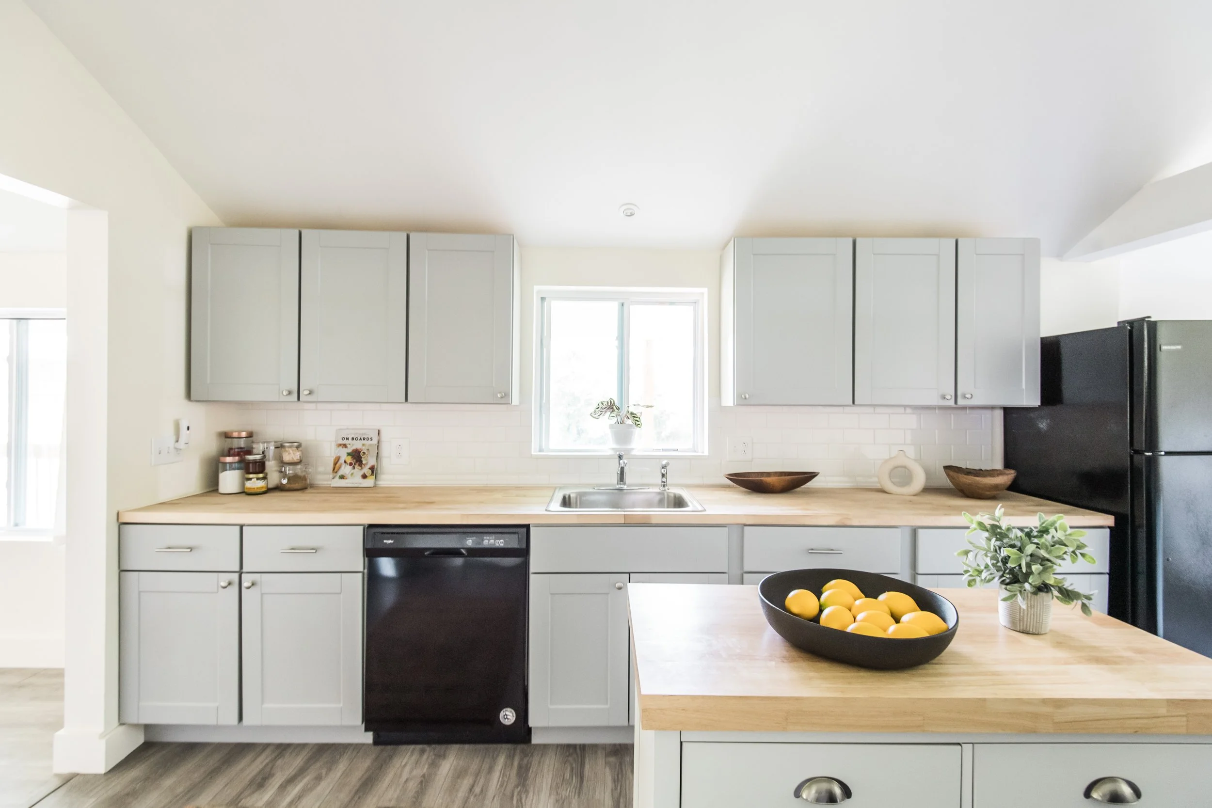 Modern kitchen with light gray cabinets, a black dishwasher, and a black refrigerator. The countertop has a bowl of lemons and a small potted plant. A window above the sink brings in natural light.