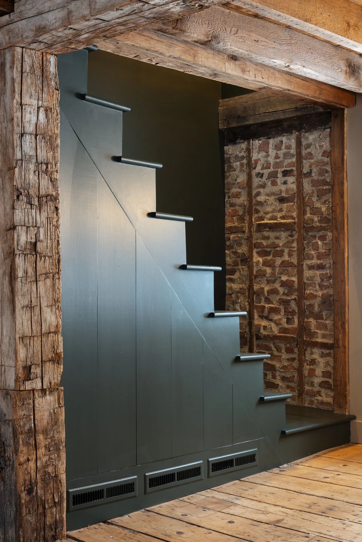 Close-up view of a modern staircase with metallic risers and wooden steps, set against exposed brick walls and a wooden ceiling, in an interior space with wooden flooring.