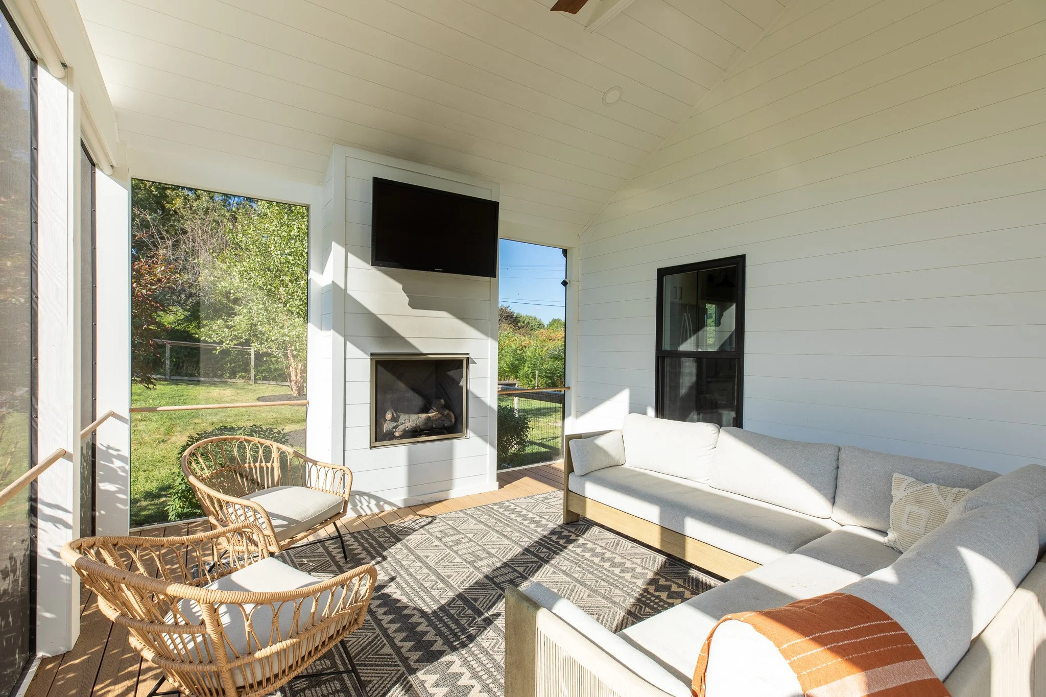 Sunlit screened porch with white sofas, rattan chairs, a patterned rug, a wall-mounted TV, and a fireplace, overlooking a green yard.
