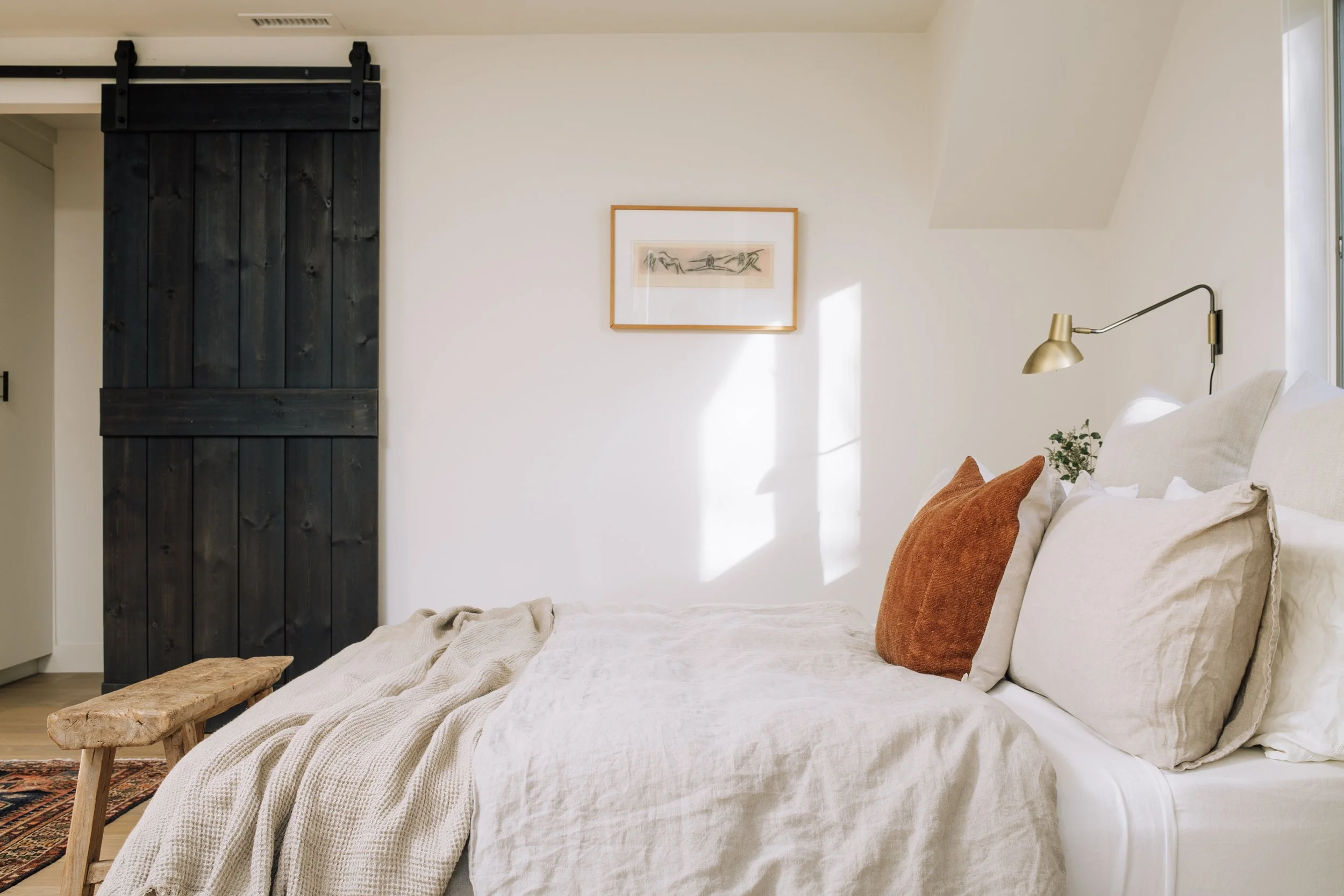 A cozy bedroom with a white bed, beige and rust pillows, a black sliding barn door, a small wooden bench at the foot of the bed, a picture frame on the wall, and a gold adjustable wall lamp.