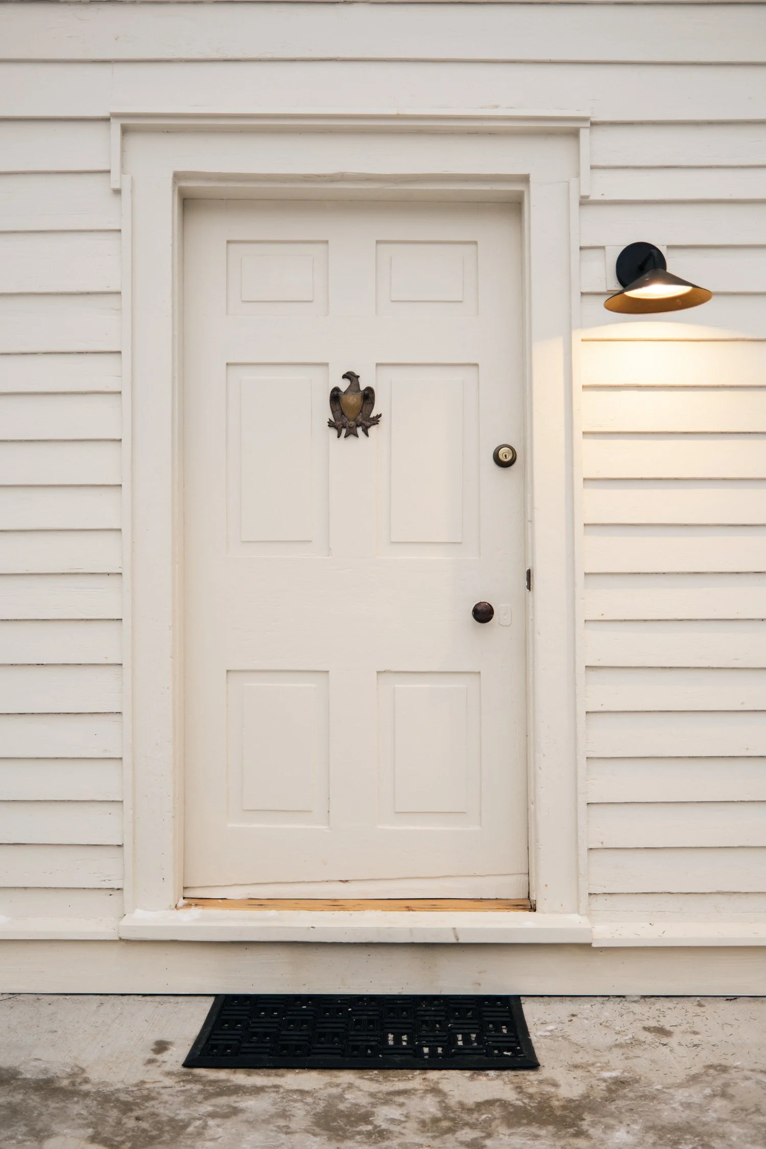 White front door with a bronze eagle door knocker, black doorknob, and a black wall-mounted light fixture on a white wooden house exterior with horizontal siding.