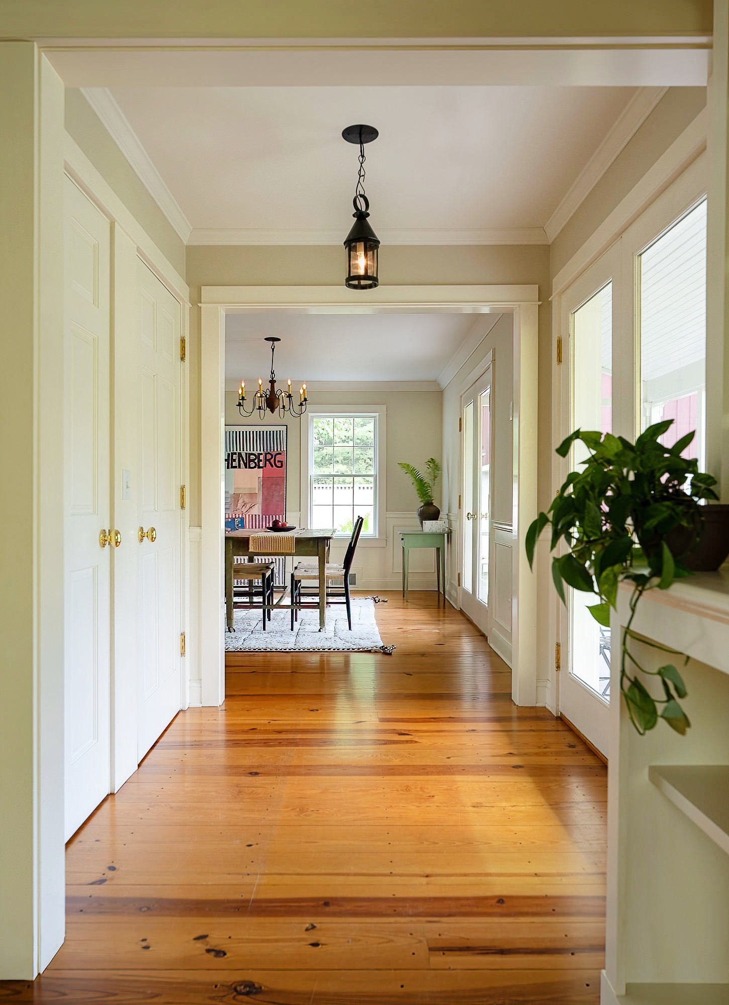 Bright hallway leading to a dining area with wooden floors, white walls, large windows, a chandelier, and plants.