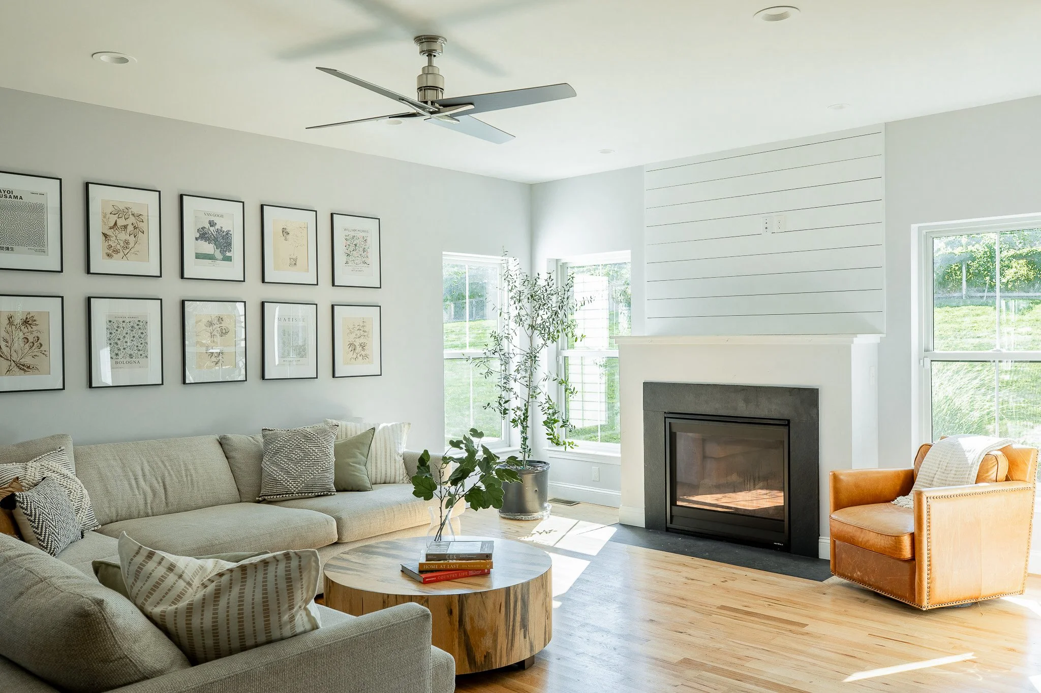 Bright living room with a beige sectional sofa, a round wooden coffee table, and an orange armchair, with white walls, framed artwork, a fireplace, and large windows showing a grassy yard.