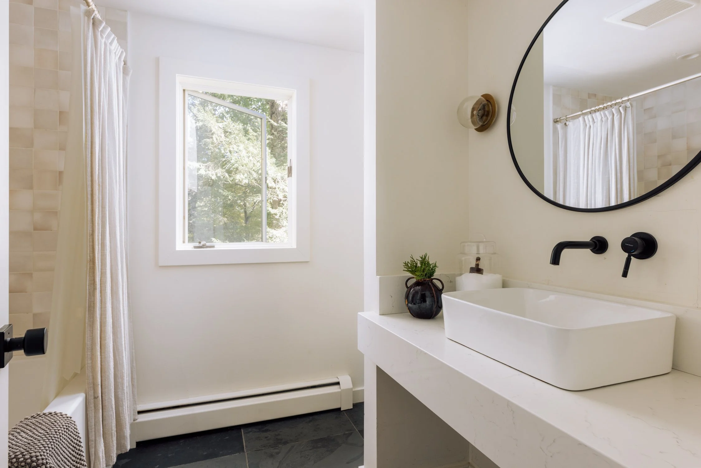 Bathroom with a white vanity, rectangular sink, round mirror, black faucet, small potted plant, and a window with white curtains and trees outside.