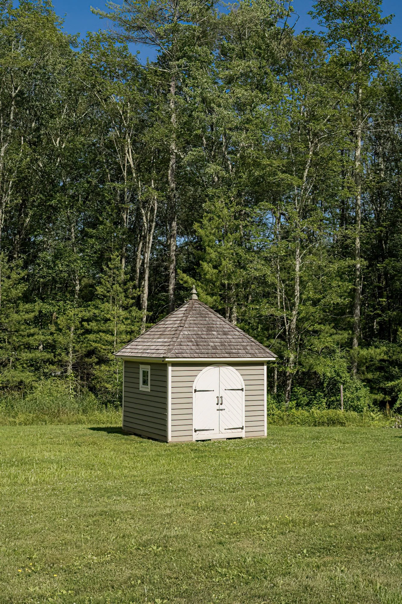 A small, beige storage shed with a shingled roof and white double doors is situated on a grassy lawn with a background of tall green trees under a clear blue sky.